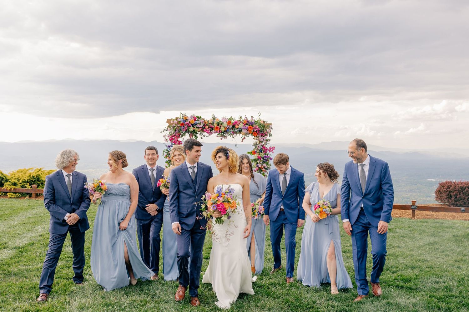 Wedding party in blue attire walking together on a grassy hilltop with a colorful floral arch behind them.