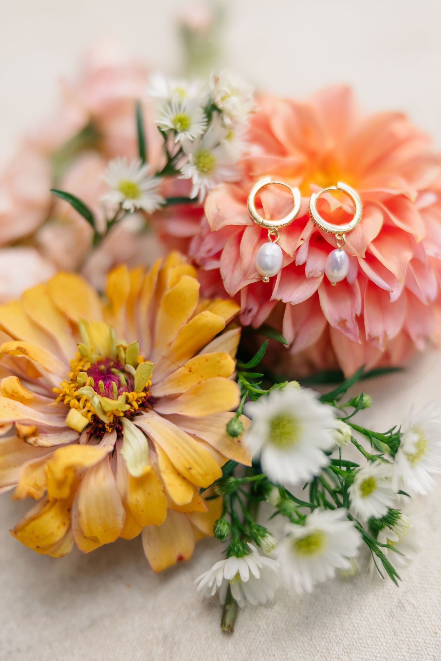 Gold hoop earrings with pearl drops resting on pink dahlia flowers surrounded by yellow zinnia and white daisies.