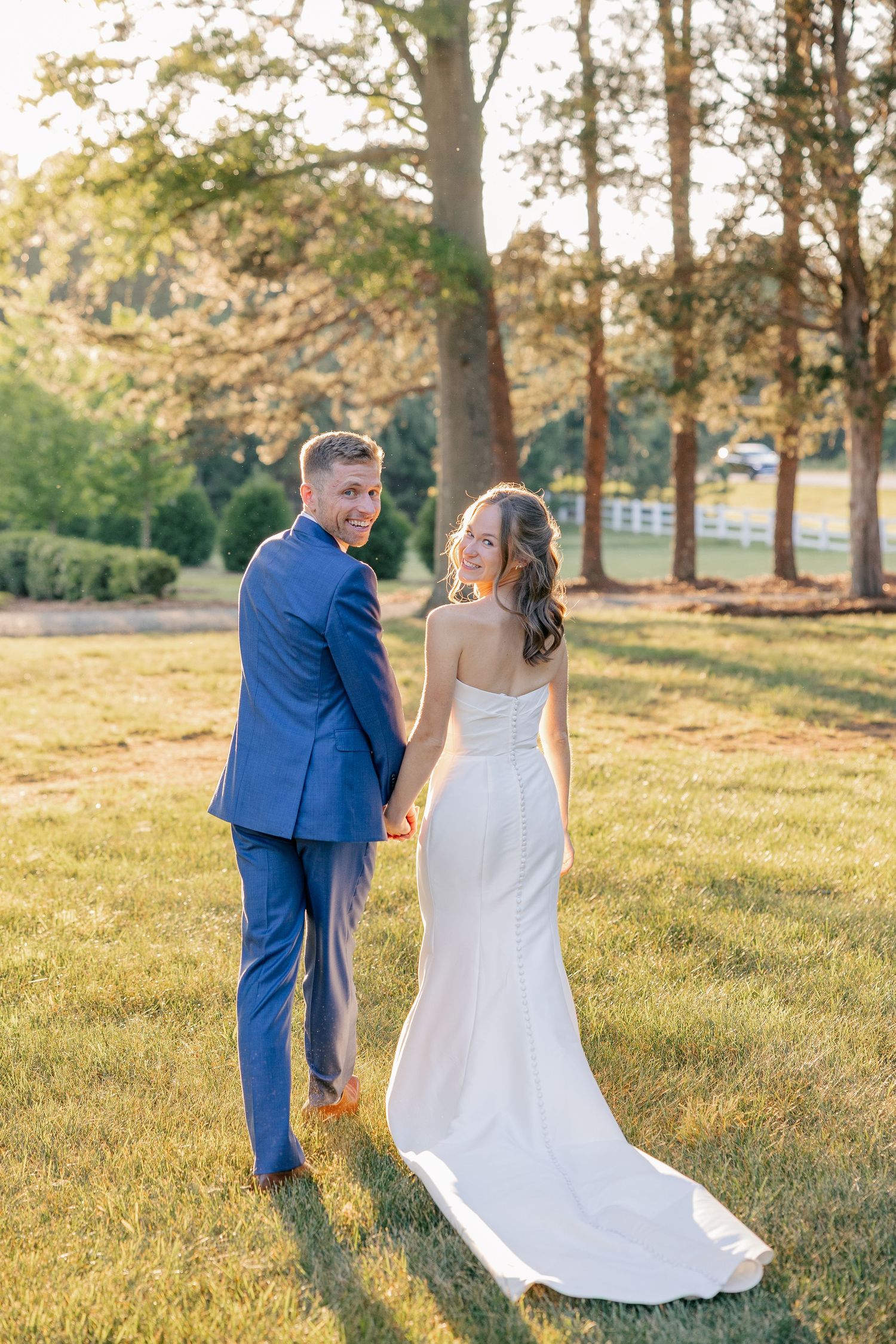 Bride and groom holding hands walking through lush green lawn at golden hour wedding.