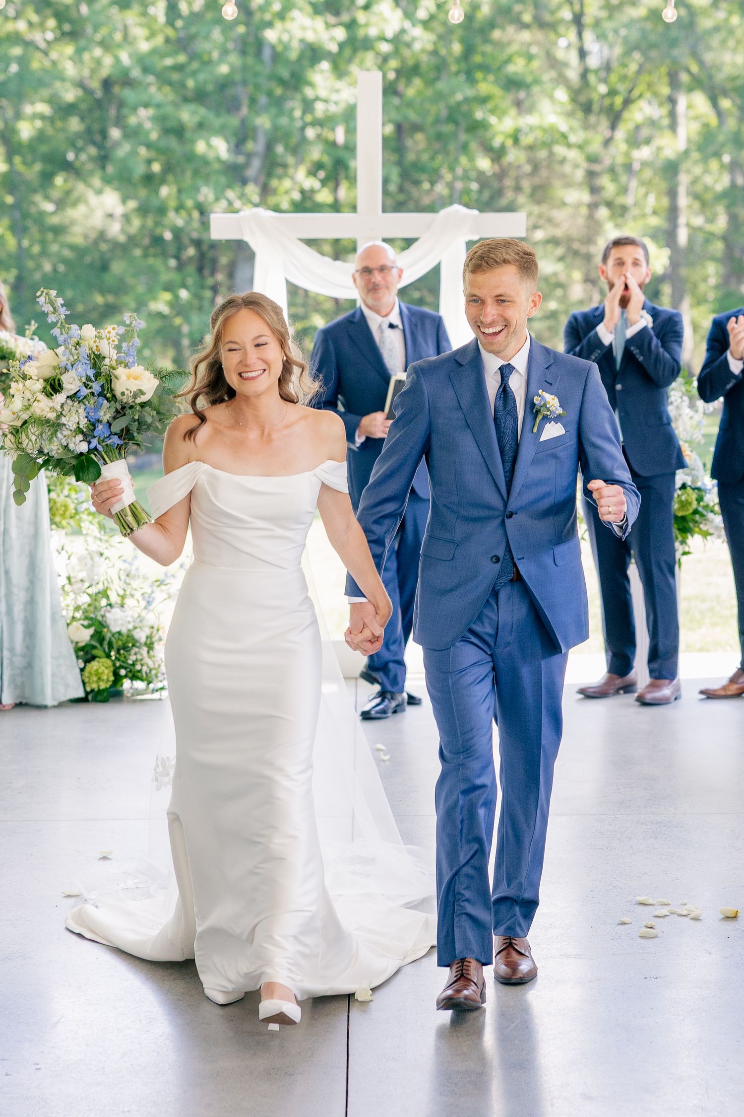 Joyful bride and groom walk back down aisle after wedding ceremony outdoors with wedding party behind them.