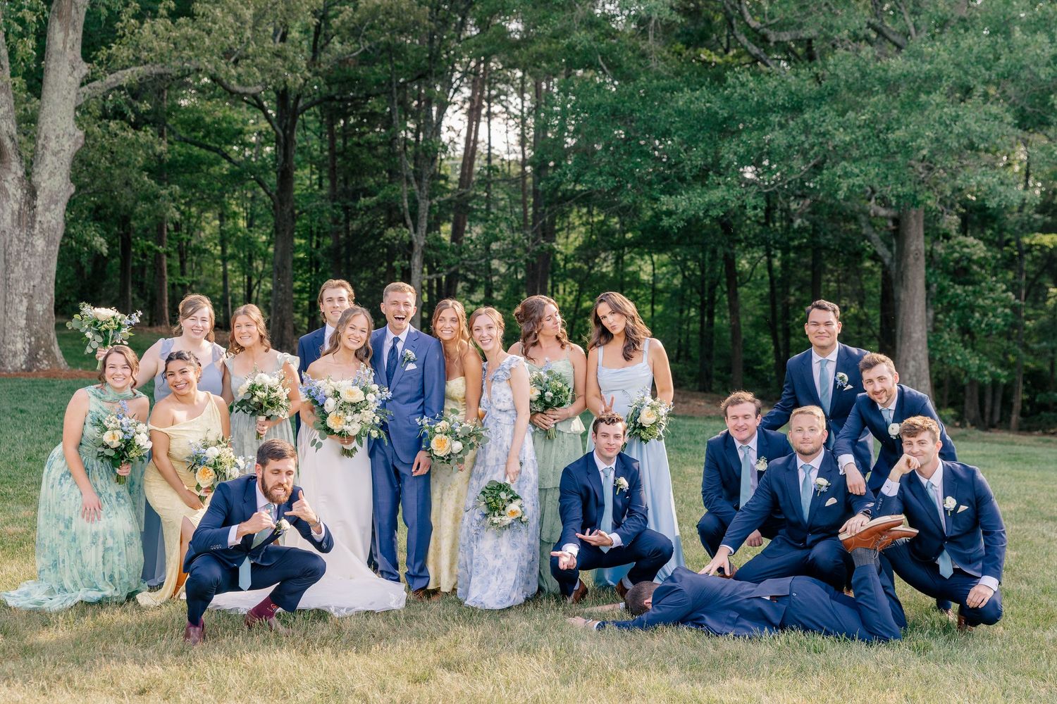 Wedding party poses together outdoors in navy and pastel attire with lush green trees behind.