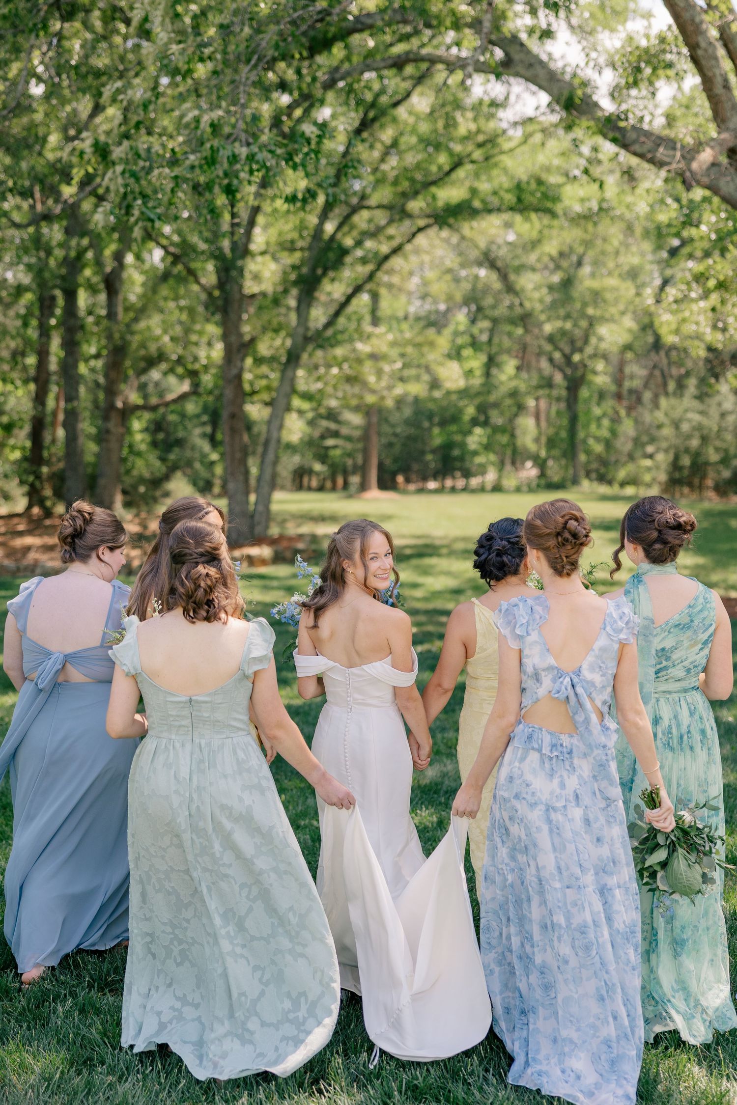 Bride in white gown walks with bridesmaids in blue and green floral dresses through lush green outdoor setting.