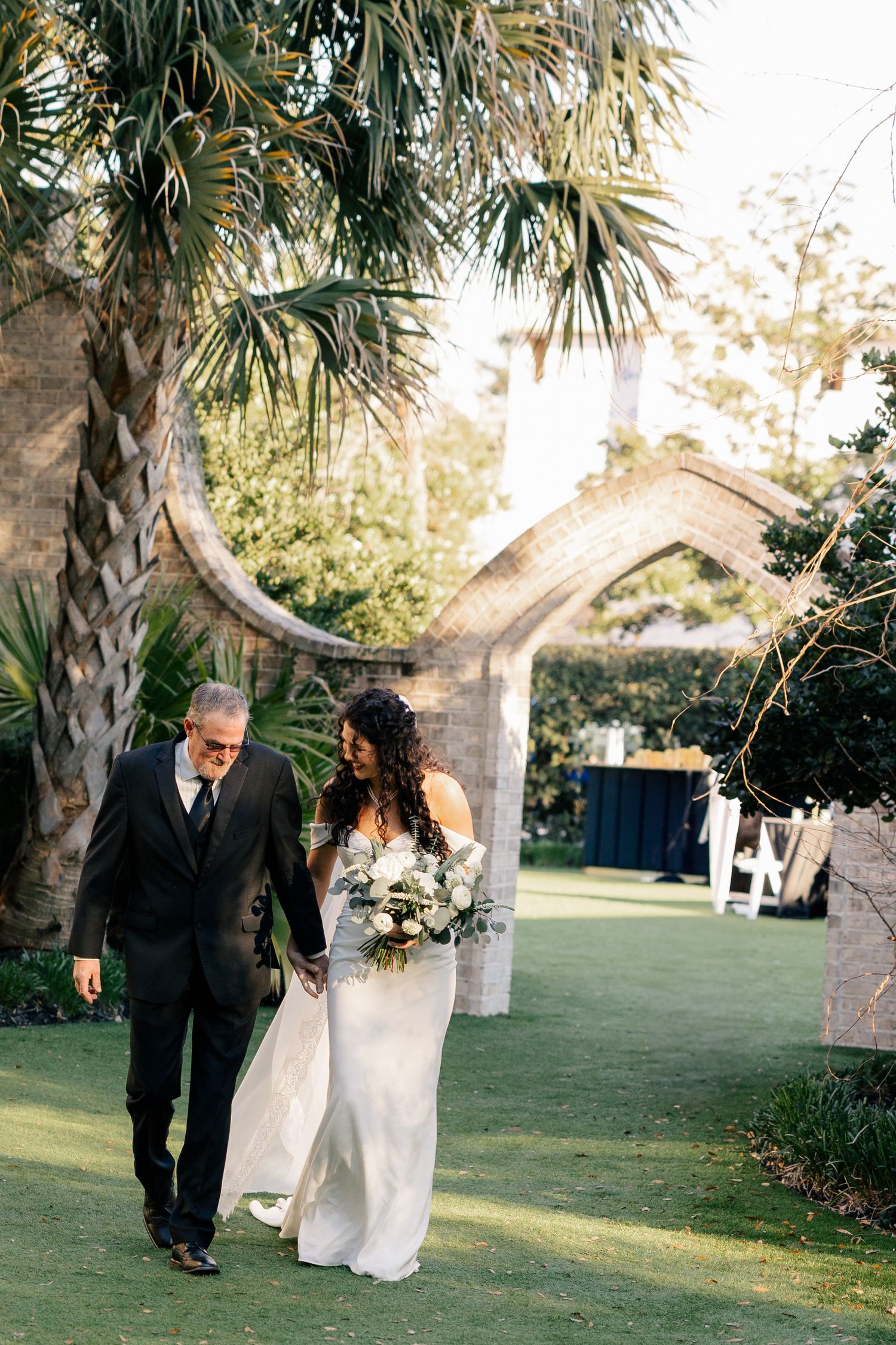 A couple walks together on a lush garden path under palm trees during an elegant outdoor wedding ceremony.