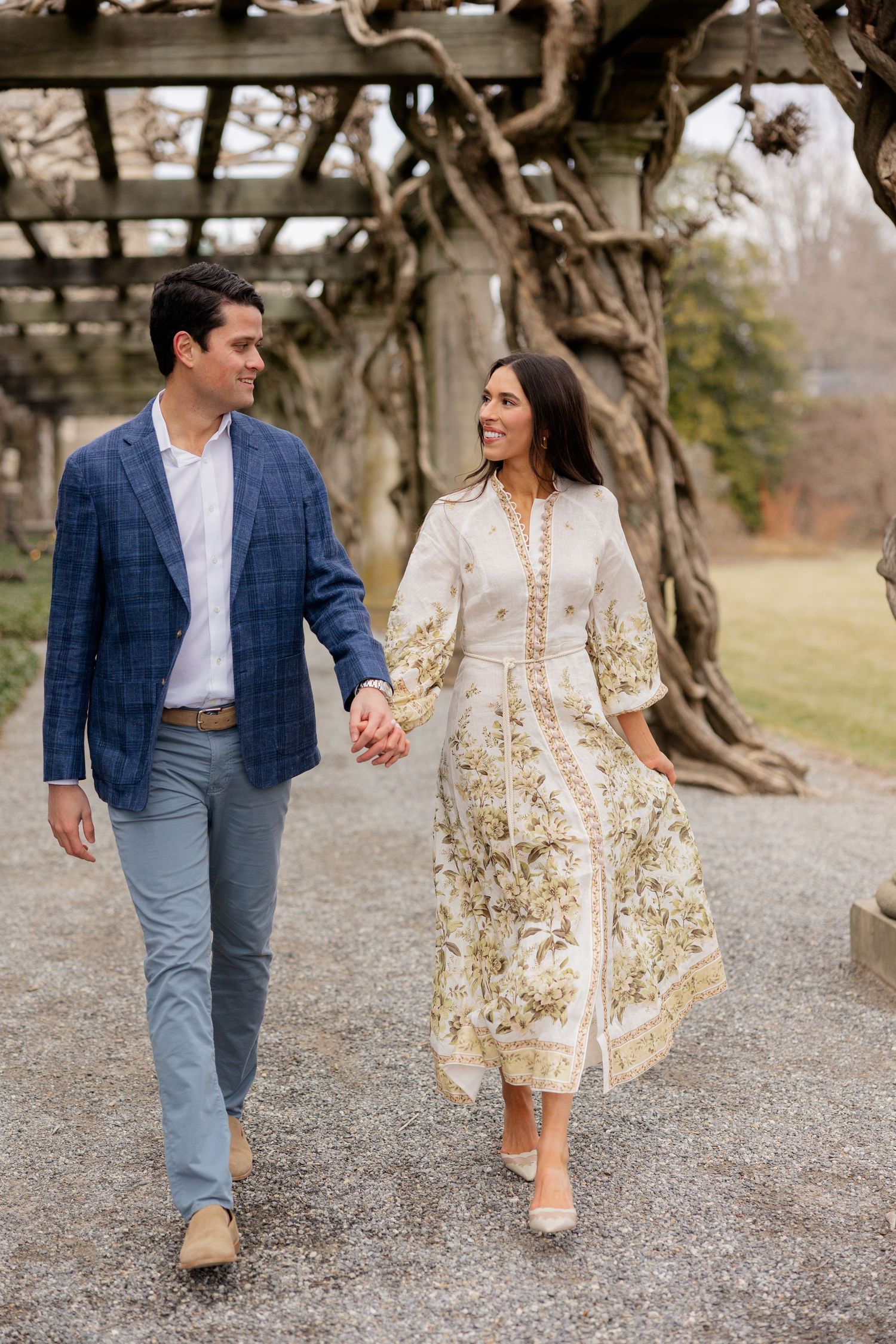 A couple walks hand in hand on a path, with him in a blue blazer and her in a flowing floral dress.