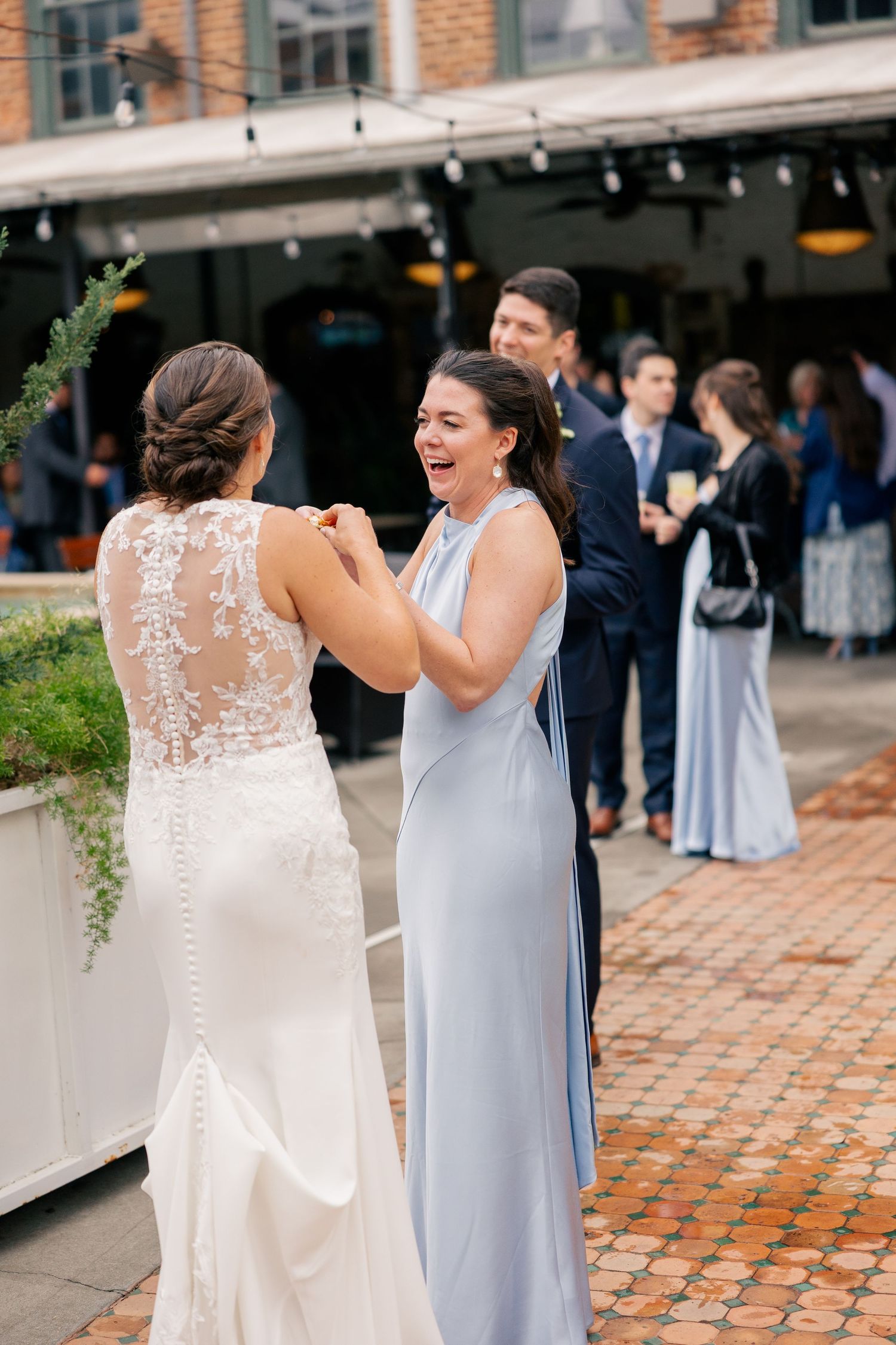 Bridesmaid in blue dress laughs joyfully with bride in lace wedding gown at outdoor wedding reception.