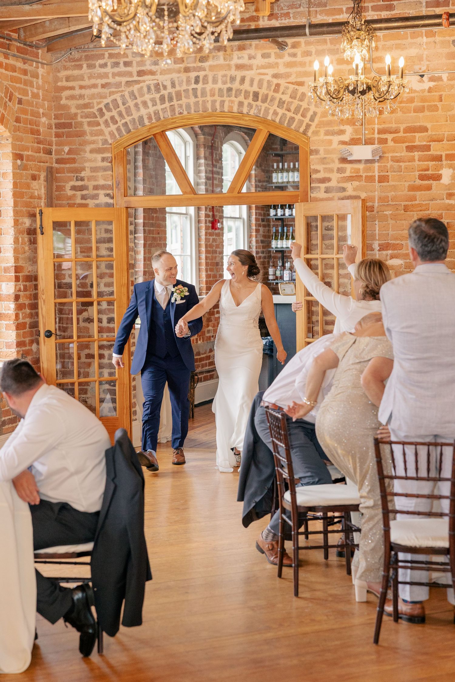 Newlyweds make their grand entrance into a brick-walled reception hall as guests cheer and celebrate.