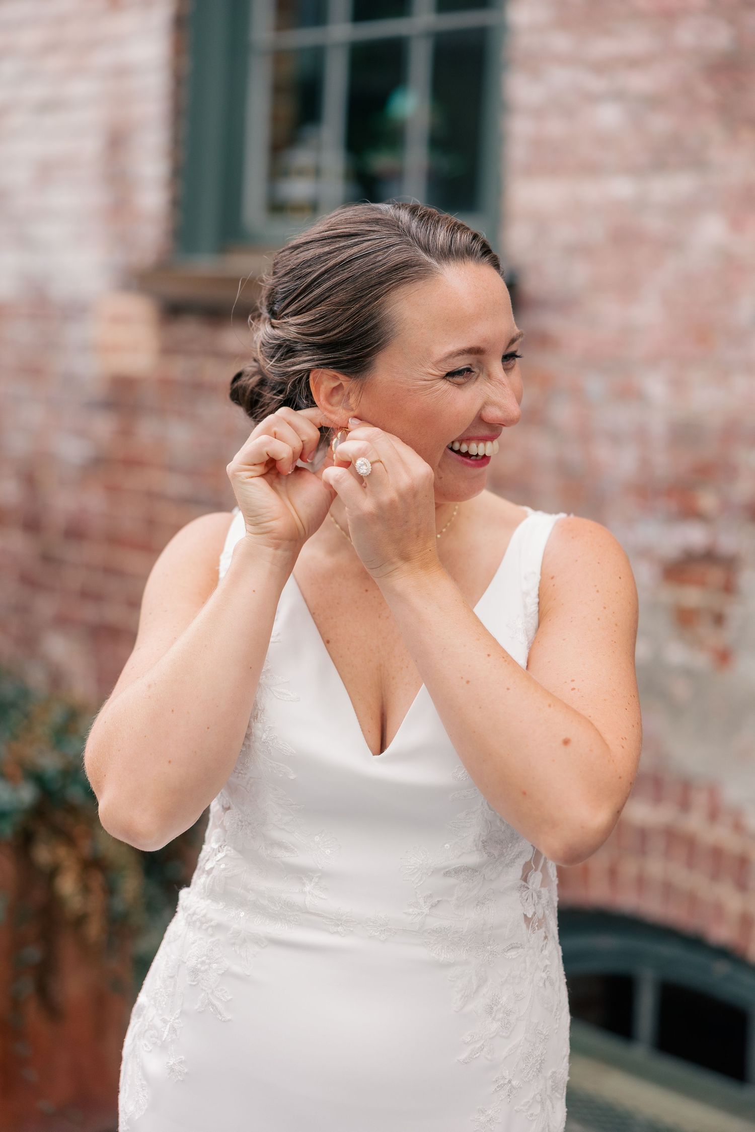 Joyful bride in white wedding dress laughing while putting on earrings against brick wall.