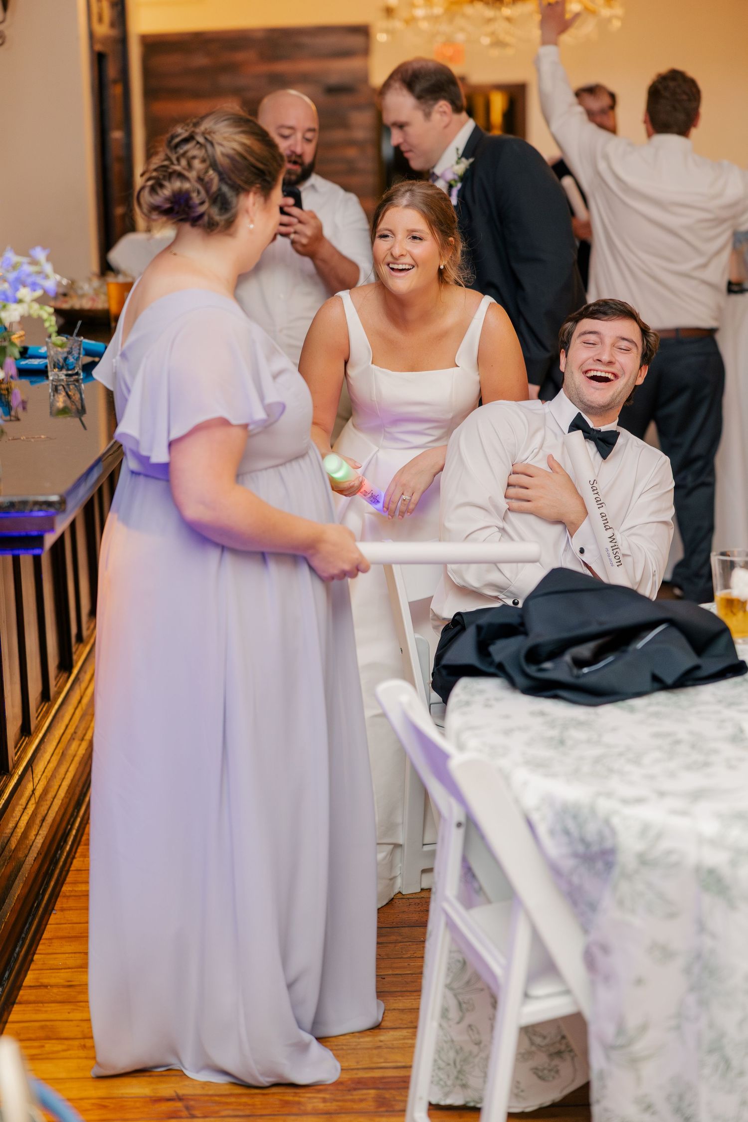 Wedding guests laugh and celebrate at reception table, bridesmaid in lavender dress holds light-up toy while others smile.