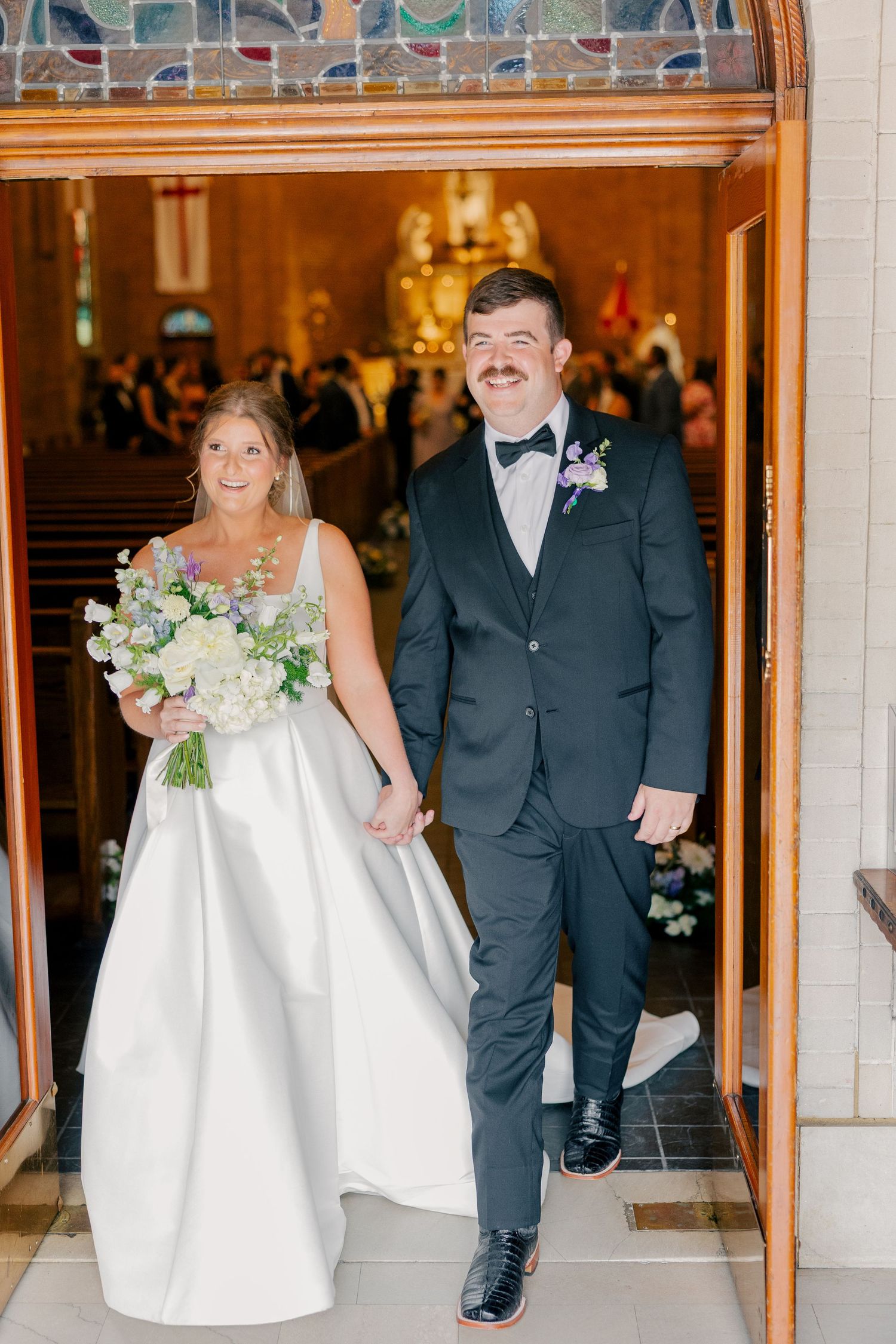 Newlyweds exit church hand in hand, bride holding white bouquet, groom smiling in black tuxedo.