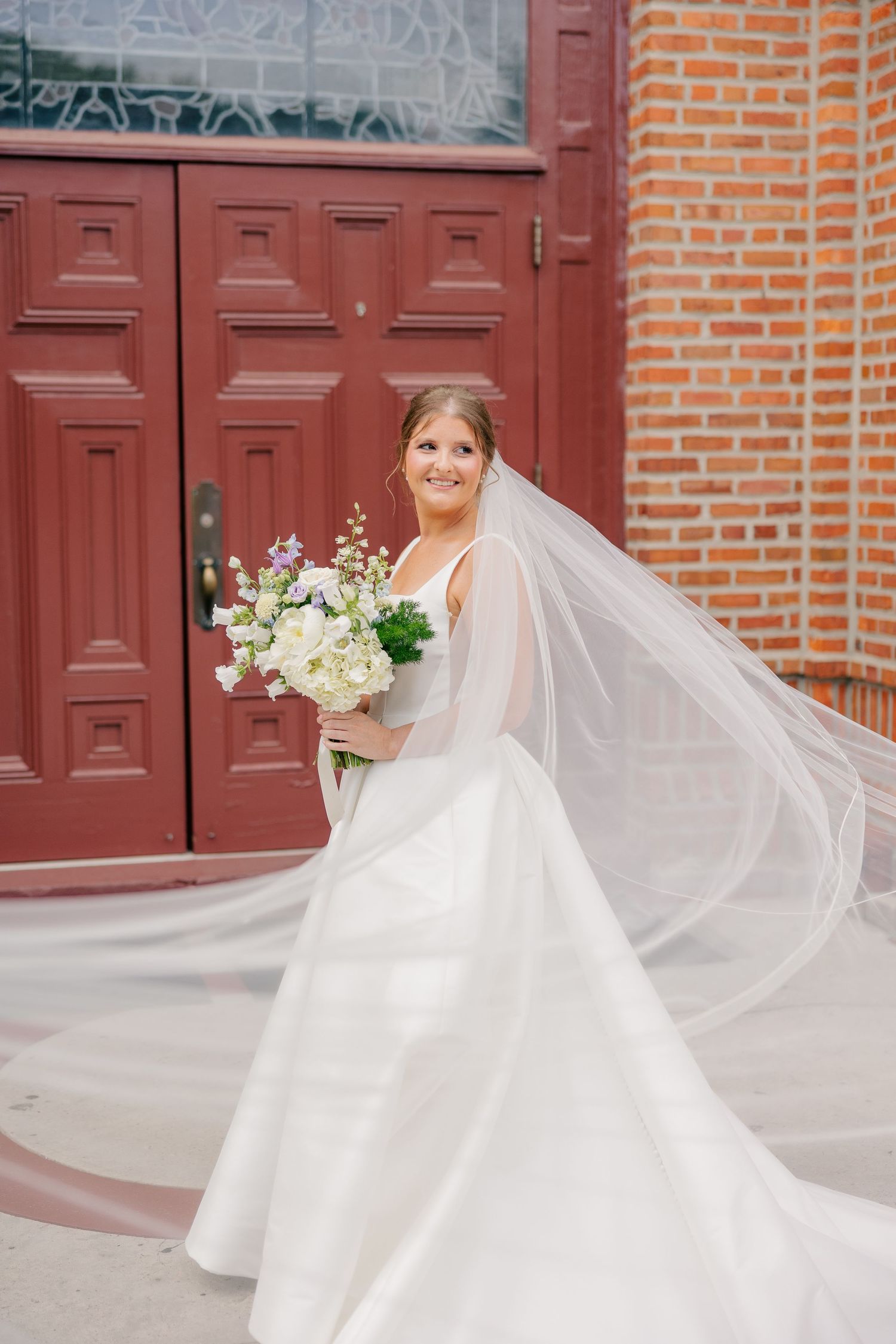 Bride twirling in elegant white ball gown with flowing veil outside church doors.