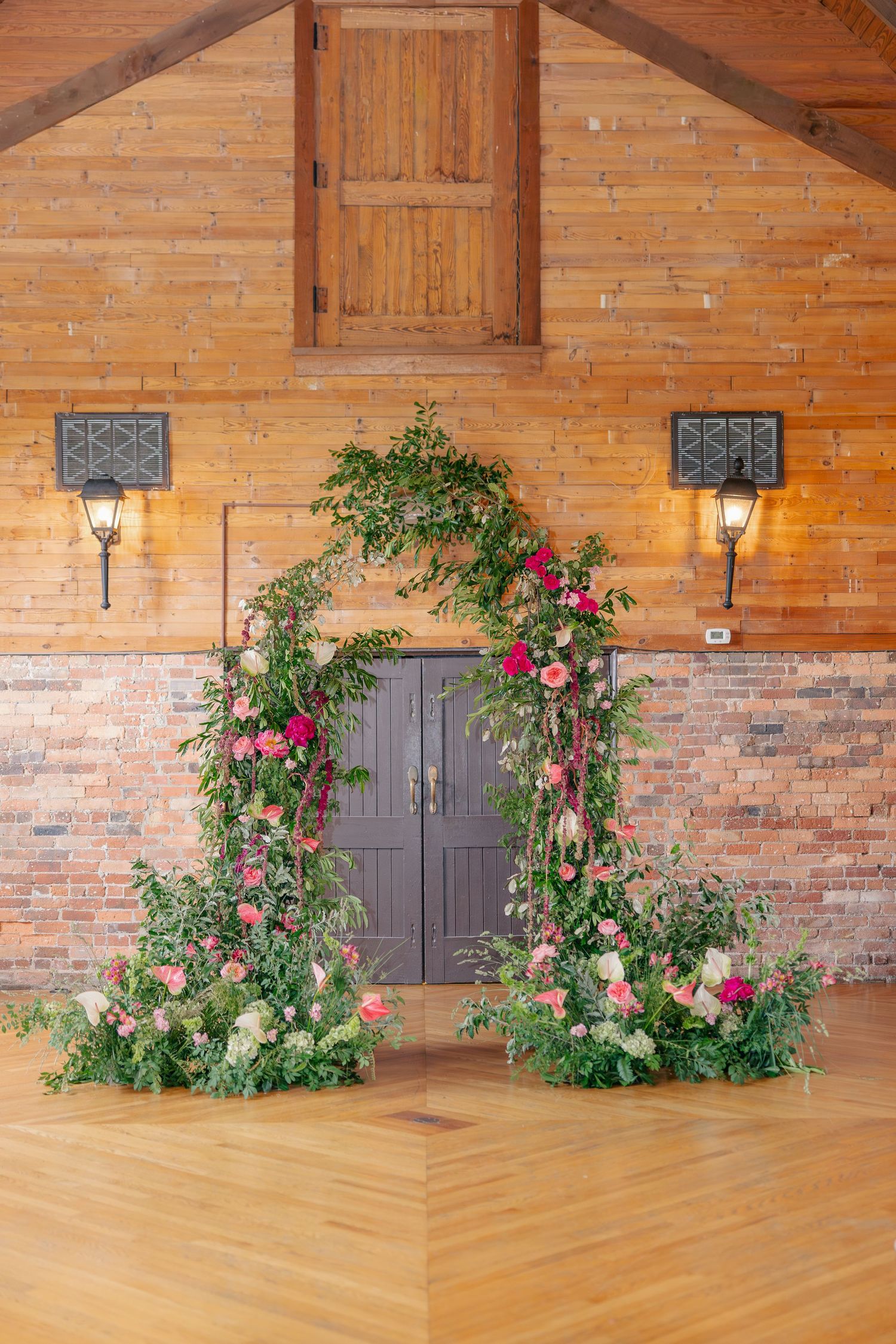Floral wedding arch with pink roses and lush greenery framing rustic barn doors on wood floors.