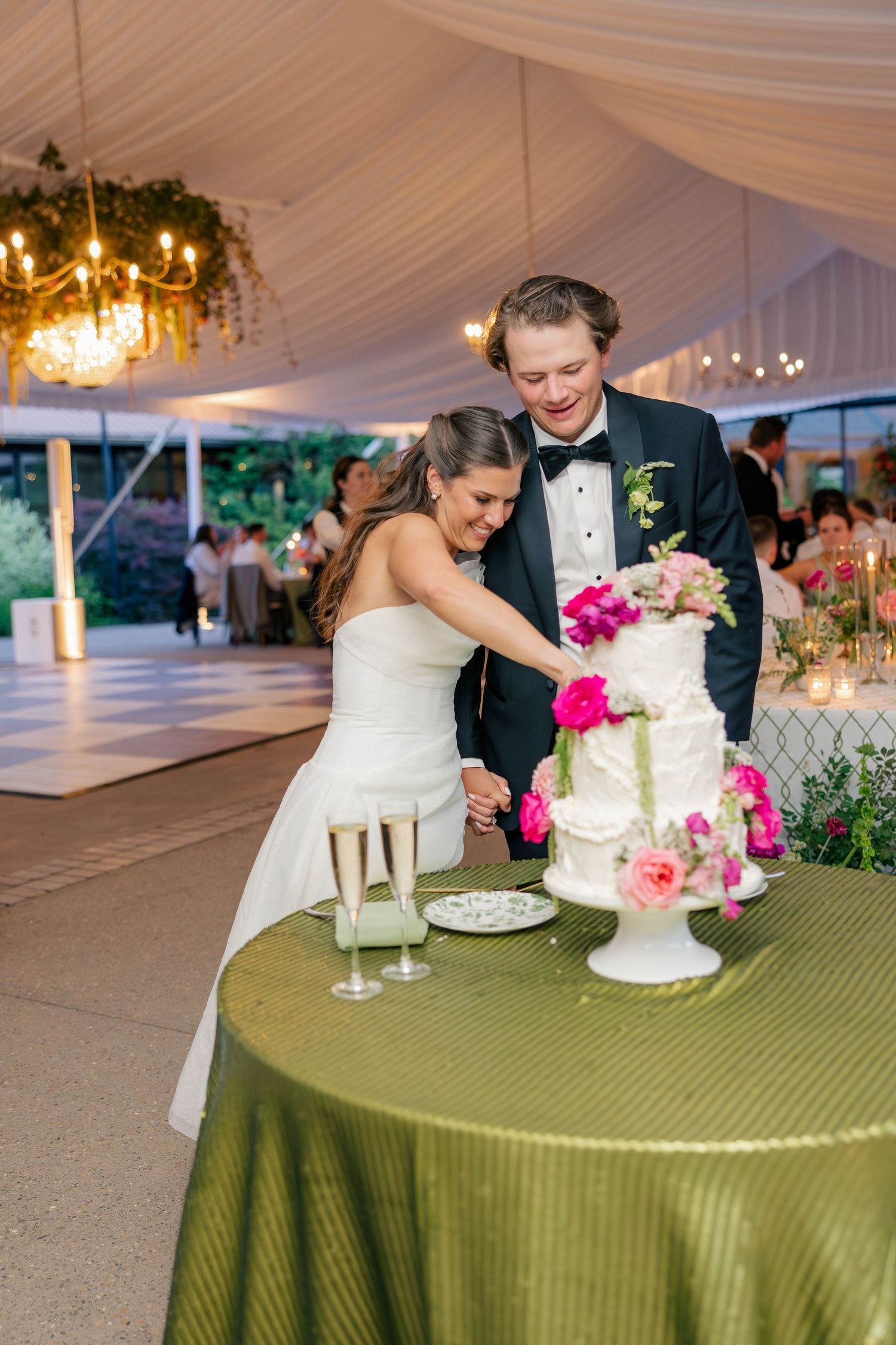 Smiling bride and groom cut elegant white wedding cake adorned with pink flowers at reception.