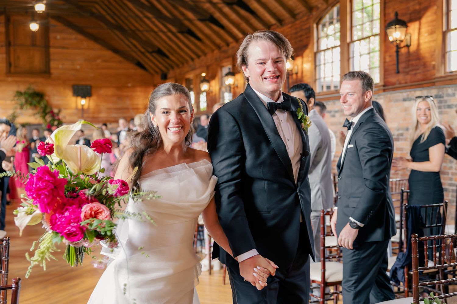Bride and groom walk down the aisle smiling, bride holding vibrant pink floral bouquet in elegant wood-beamed wedding venue.