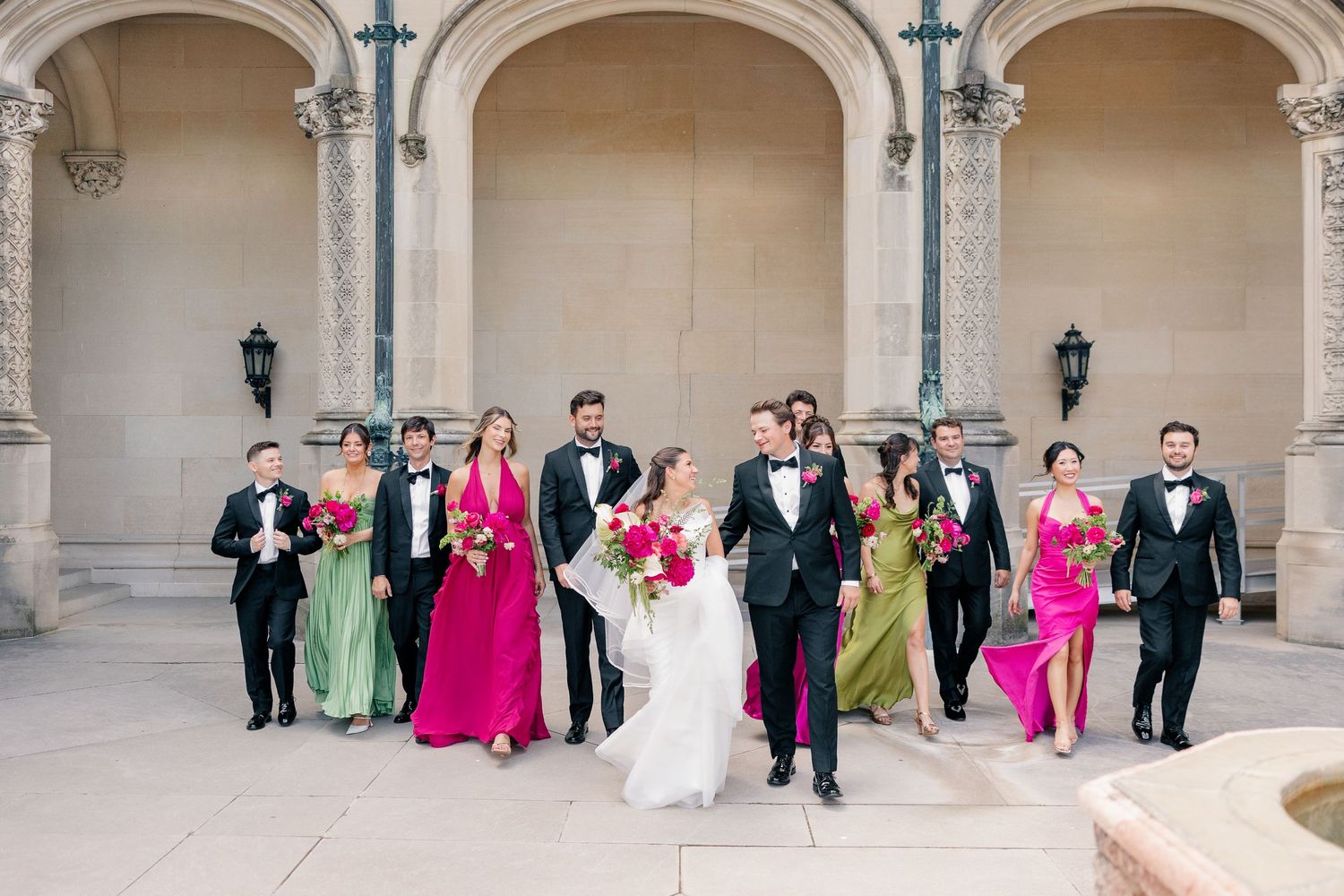 Wedding party walks together in colorful mix of pink and green gowns with black tuxedos outdoors.