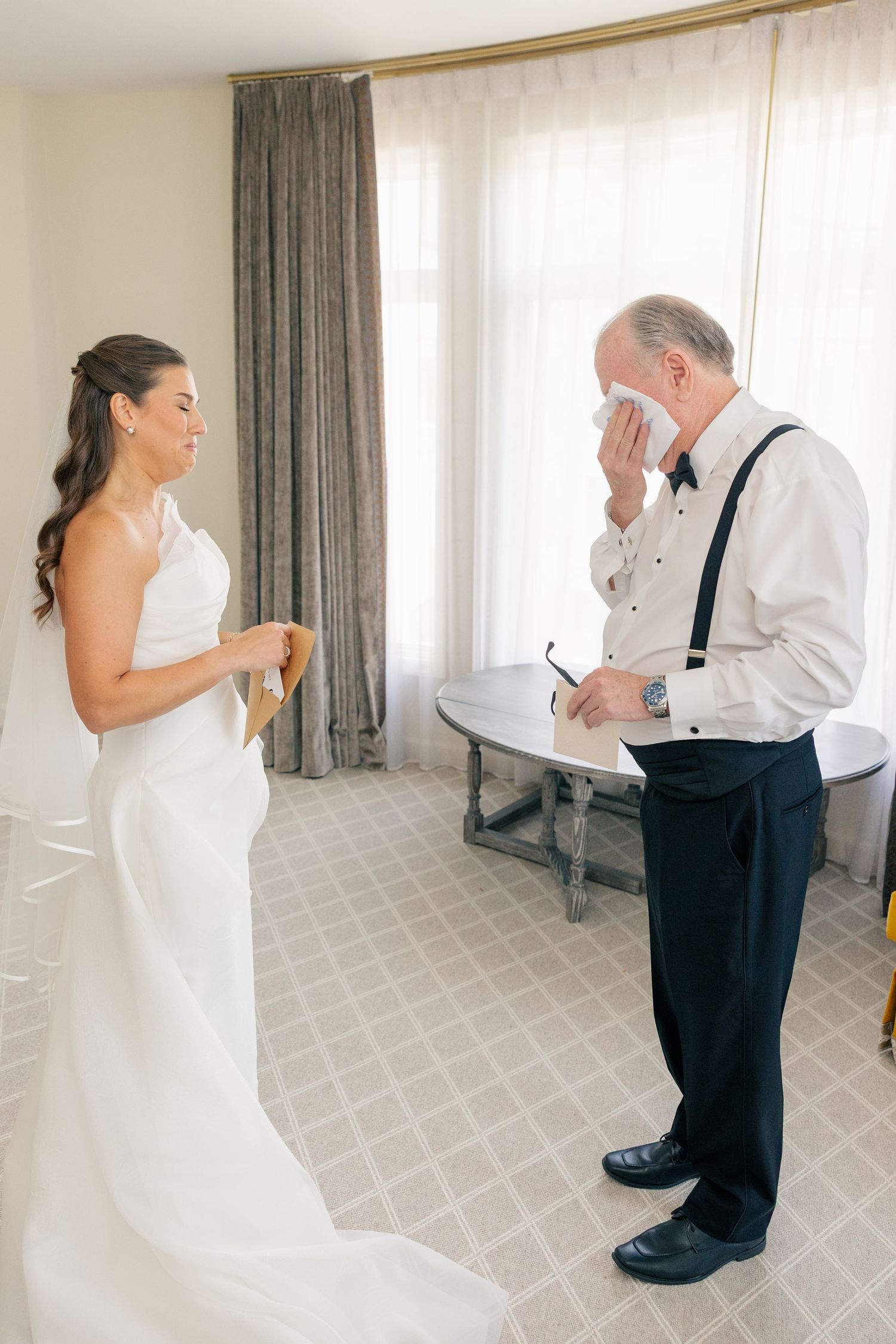 Emotional father wiping tears with handkerchief seeing bride in white wedding gown in hotel room.