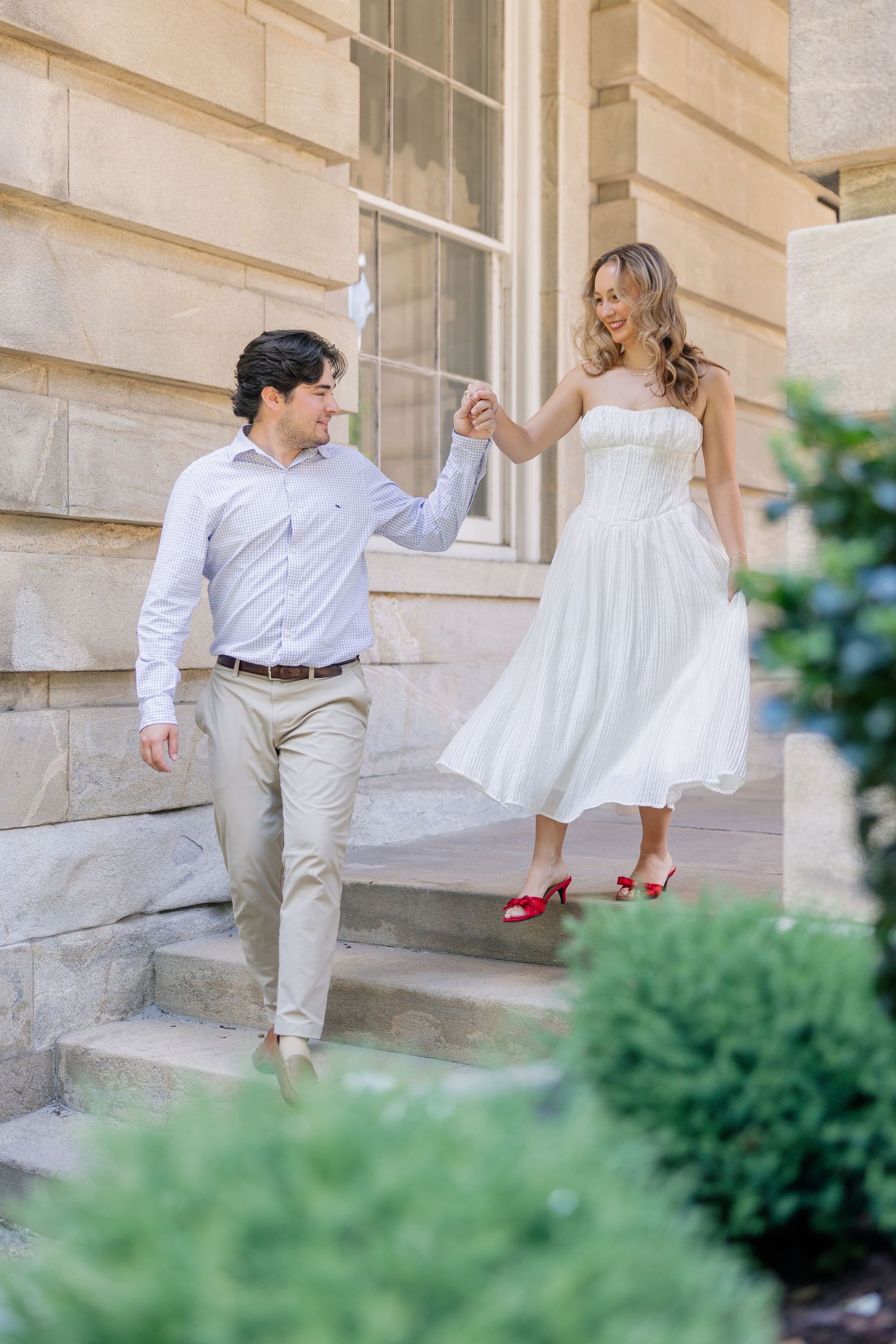 Couple holding hands walking down elegant stone steps, woman in white dress with red heels.