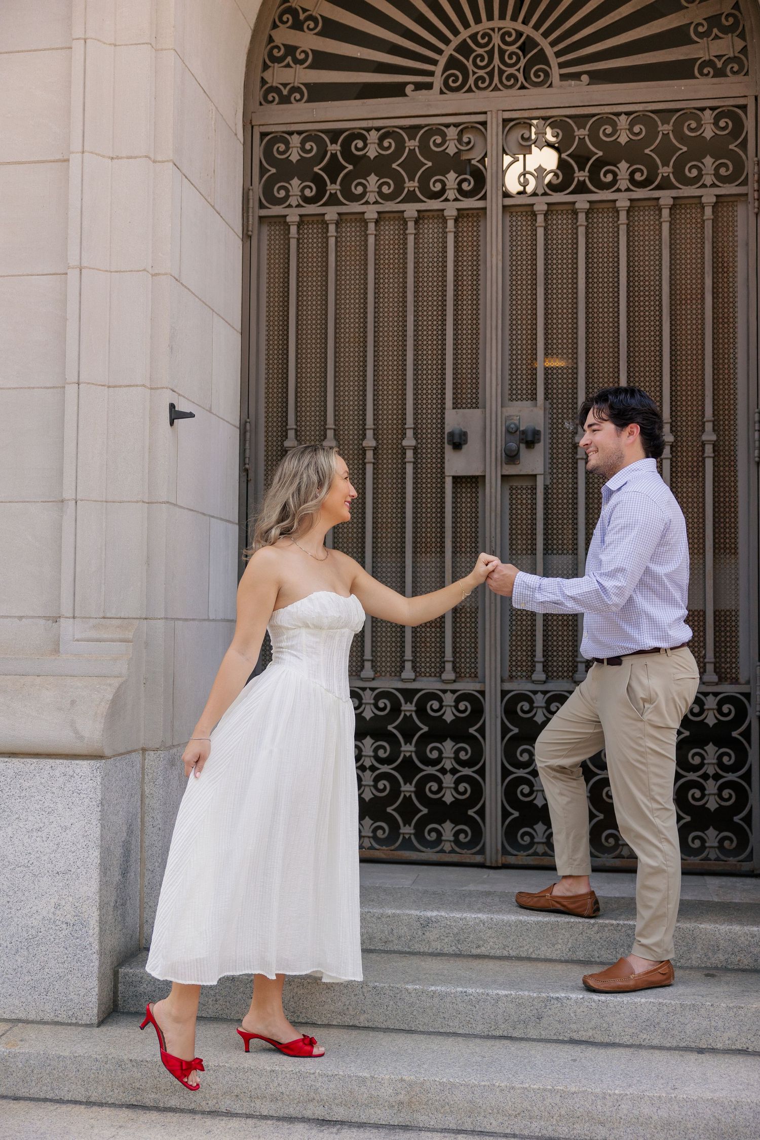 Couple holding hands on stone steps before ornate iron gate, woman in white dress with red heels, man in blue shirt.