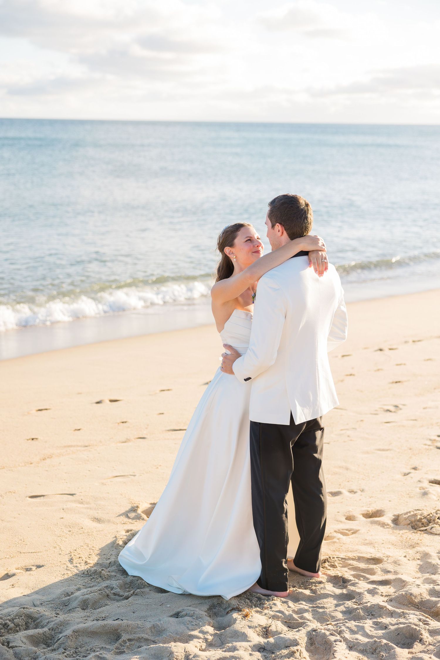 Bride and groom embrace on a sandy beach at sunset, with calm ocean waves in the background.