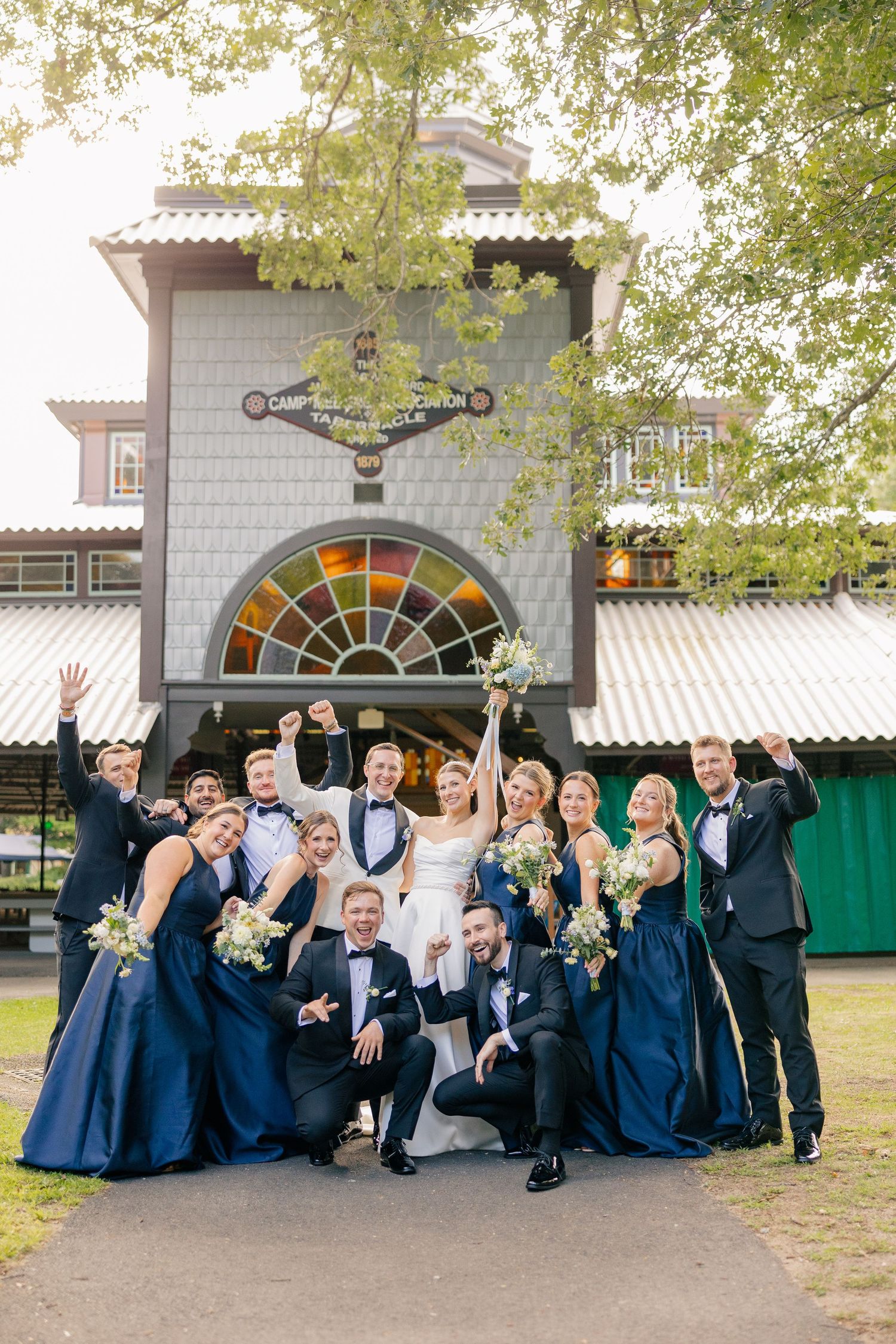 Joyful wedding party in navy and white celebrates outside a historic venue with raised arms and bouquets.