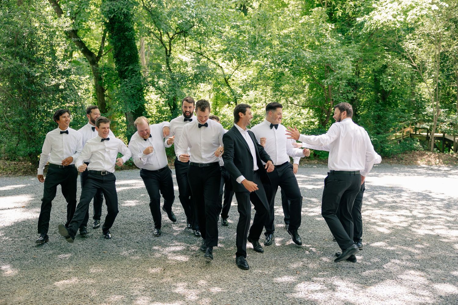 Groomsmen in black tuxedos and white shirts dance outdoors on gravel path surrounded by lush green trees.