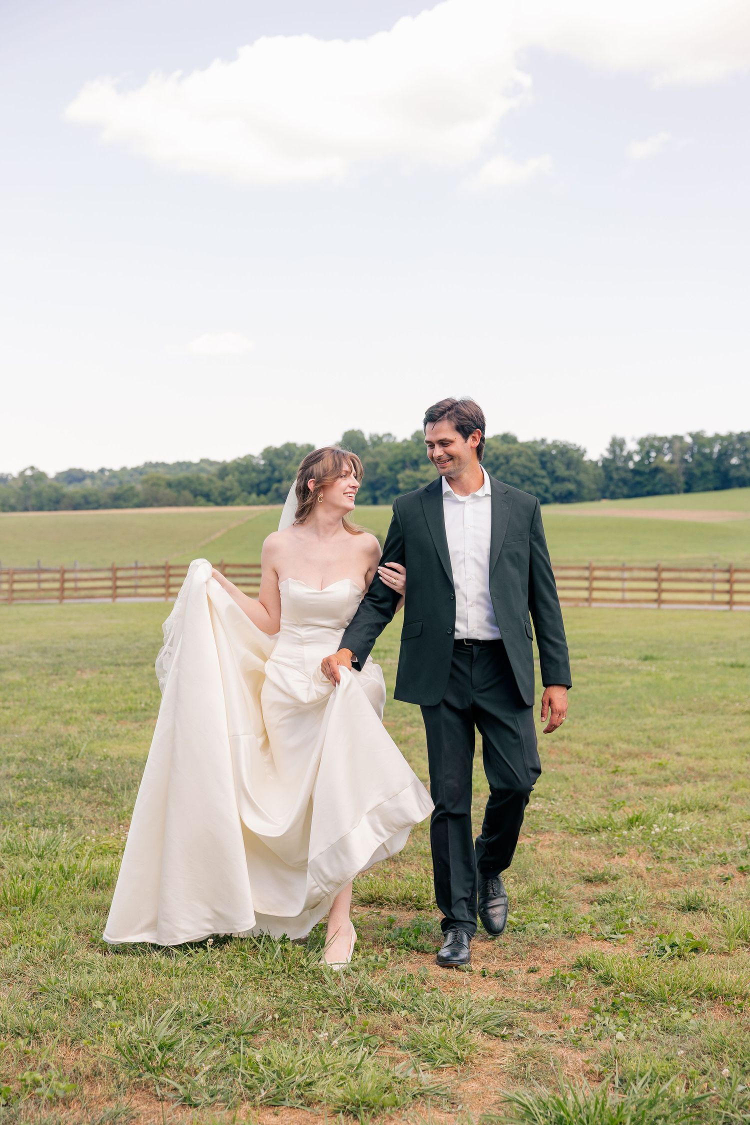 Bride in white gown and groom in black suit walking hand in hand through a green farm field.