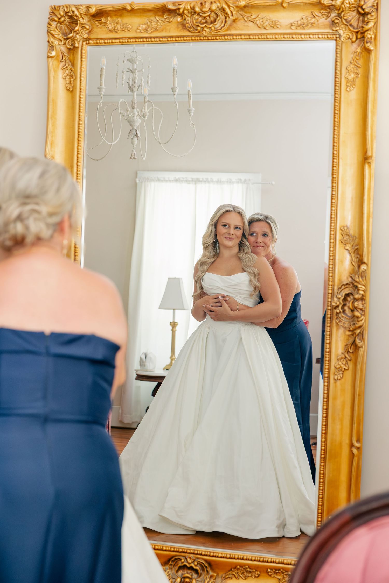 Bride in white ball gown admires her reflection in ornate gold mirror while bridesmaid in navy dress embraces her.