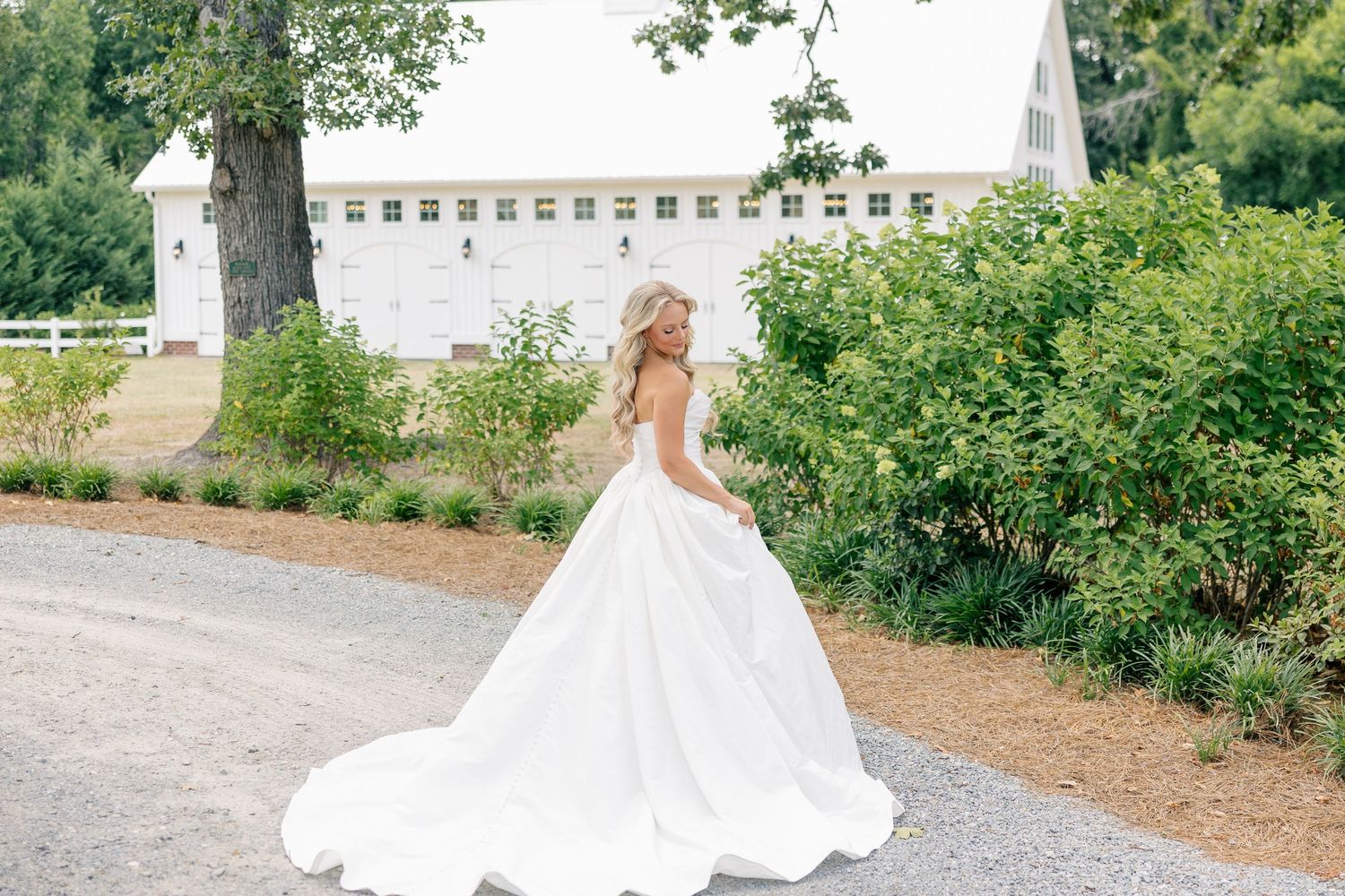 Bride in white ball gown twirling on gravel path beside white wedding barn.