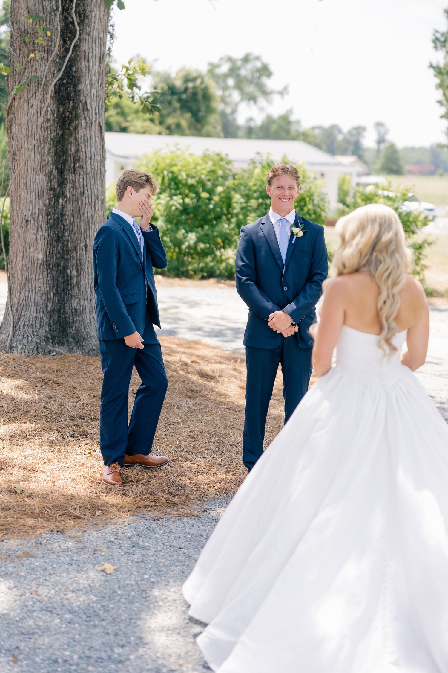 Groom smiling at bride during first look moment with groomsman beside a large tree at outdoor wedding.