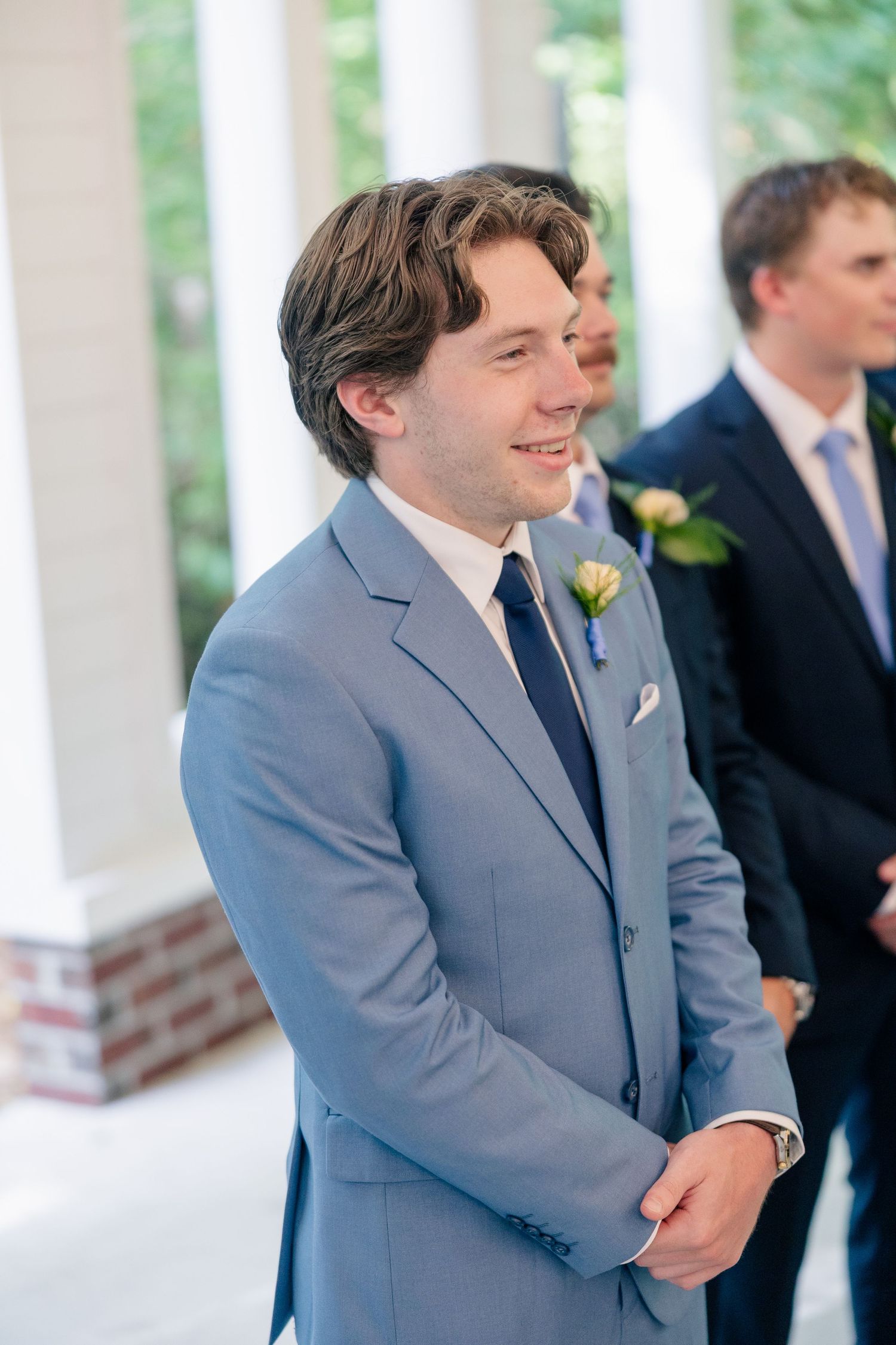 Groom in light blue suit smiling at the altar during wedding ceremony with boutonniere on lapel.