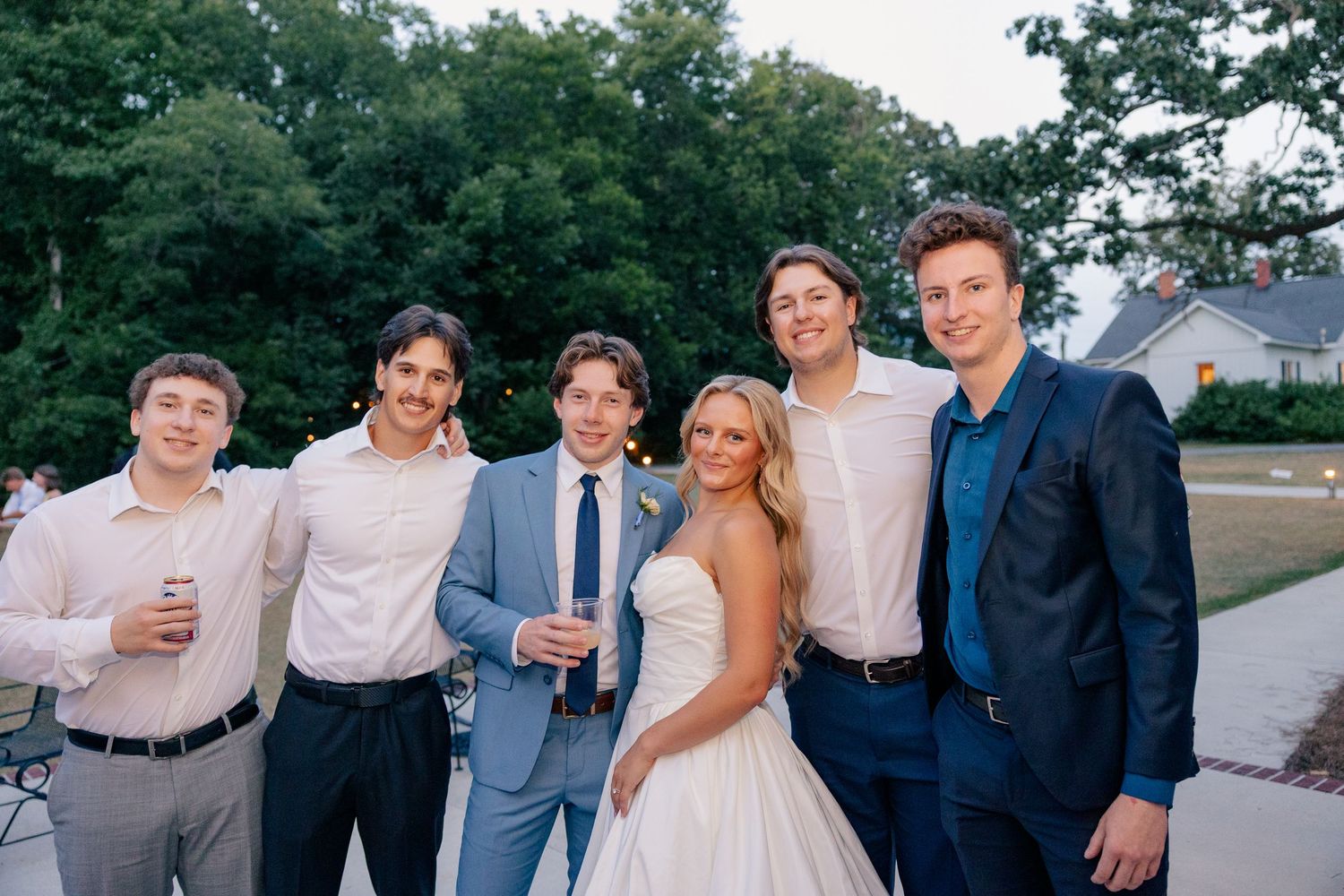 Smiling bride in white gown poses with five groomsmen in blue and pink suits at outdoor wedding.
