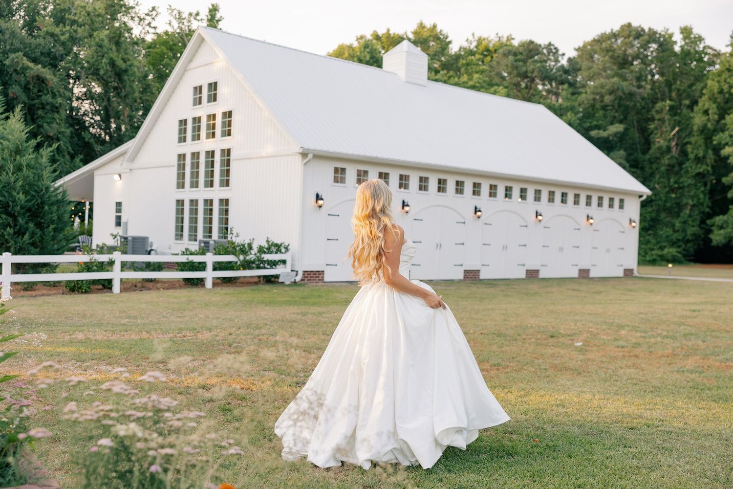 Bride in flowing white gown walks toward a charming white barn wedding venue at golden hour.