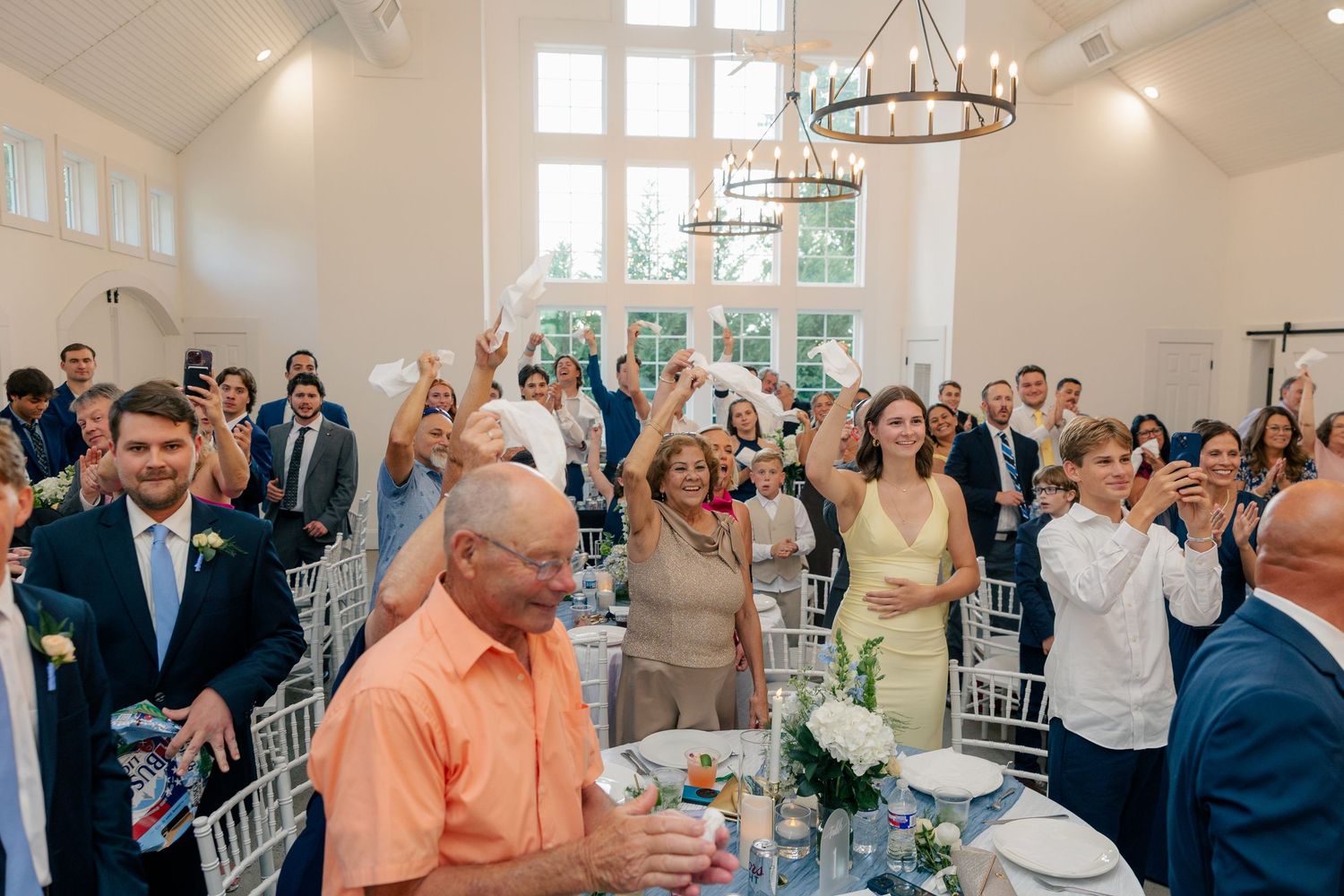Wedding reception guests cheering and raising glasses in a bright white elegant venue.