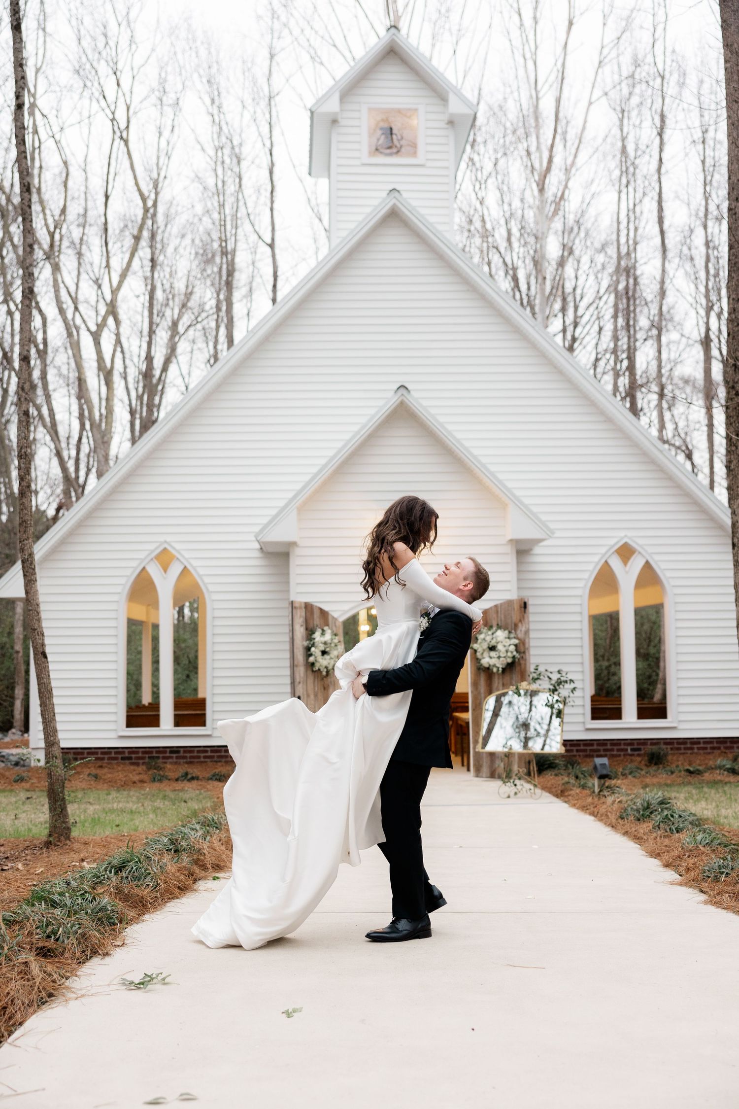 A romantic moment captured outside a charming white chapel with stained glass windows in a wooded setting.