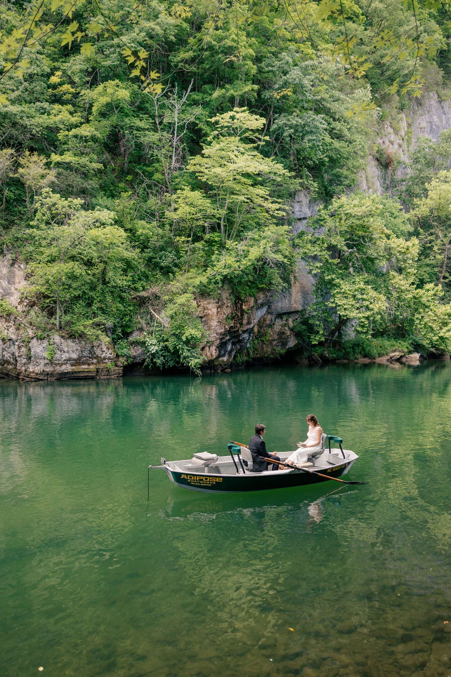 Bride and groom sitting in a small boat on a serene green river surrounded by lush forest.
