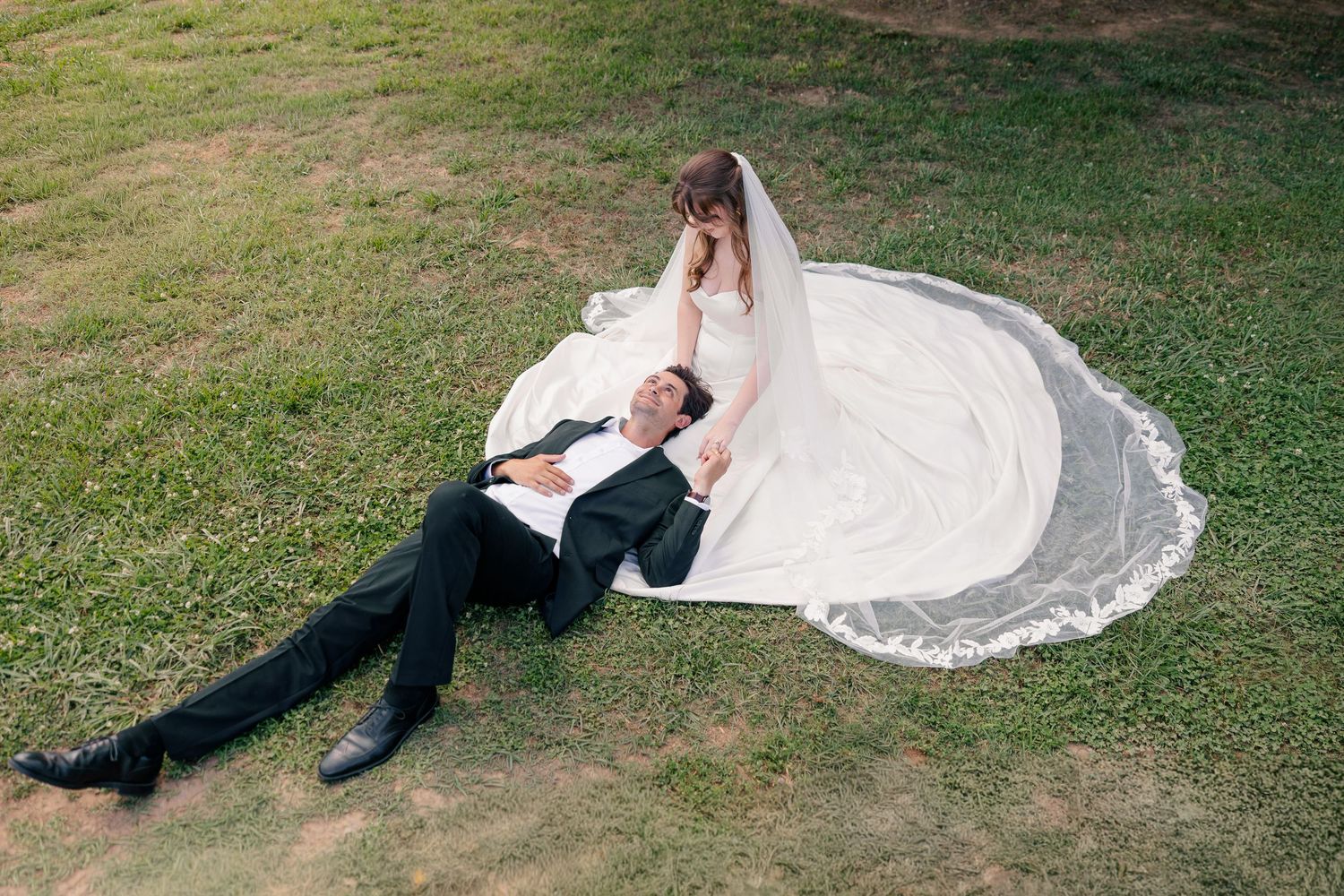 Bride and groom posing on green grass with flowing white wedding veil spread around them.