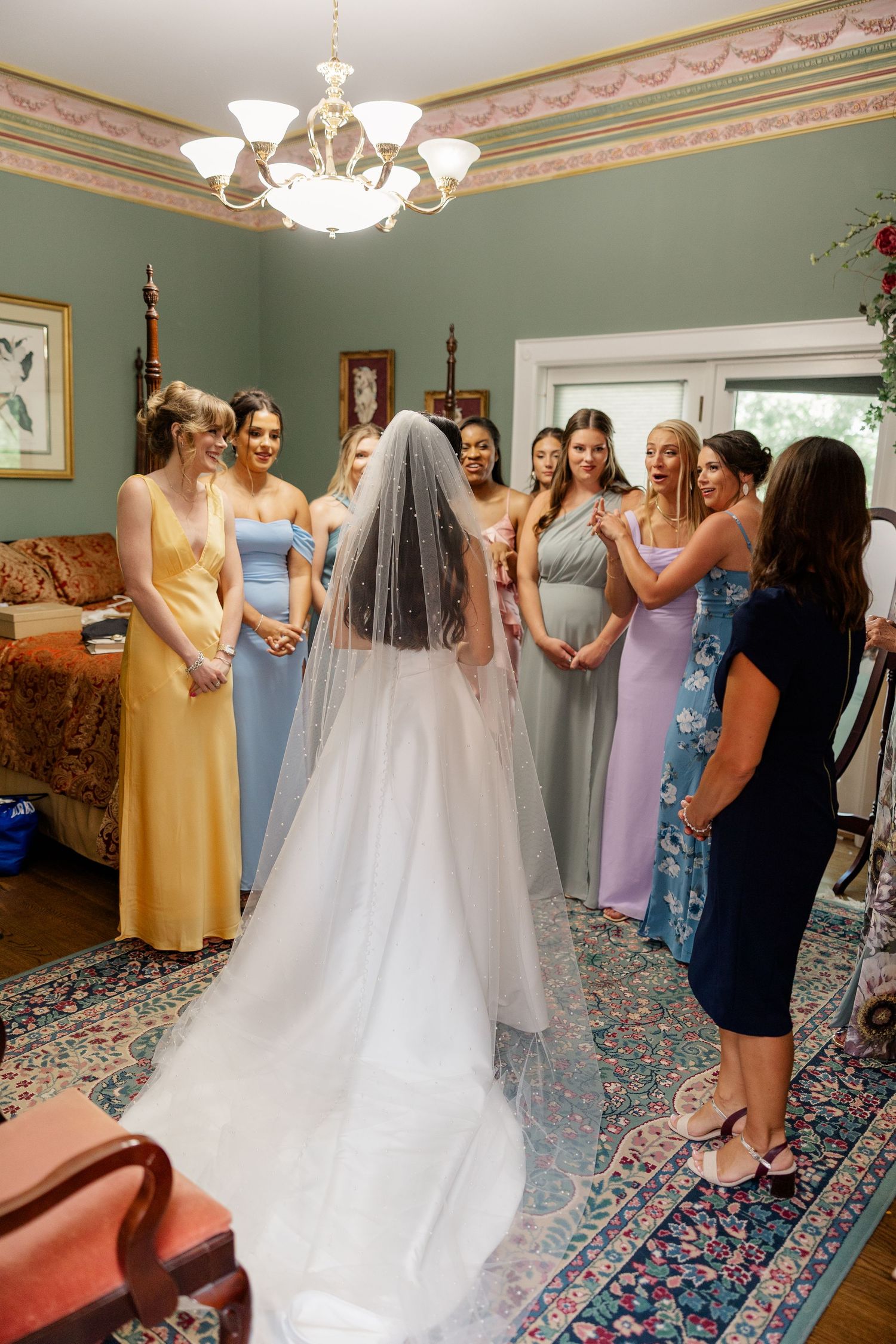 A bridal party in pastel dresses gathers in an elegant green room with a chandelier to admire the bride.