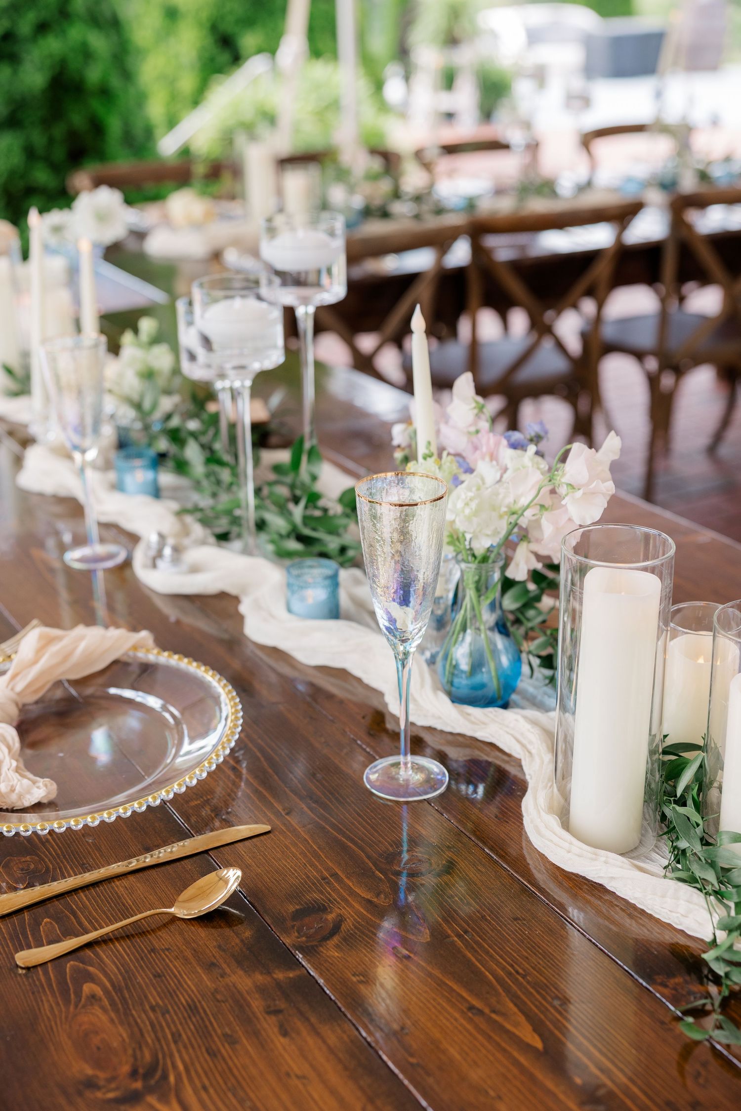 Elegant wedding table setting with blue vases, candles and greenery on a rustic wooden table.