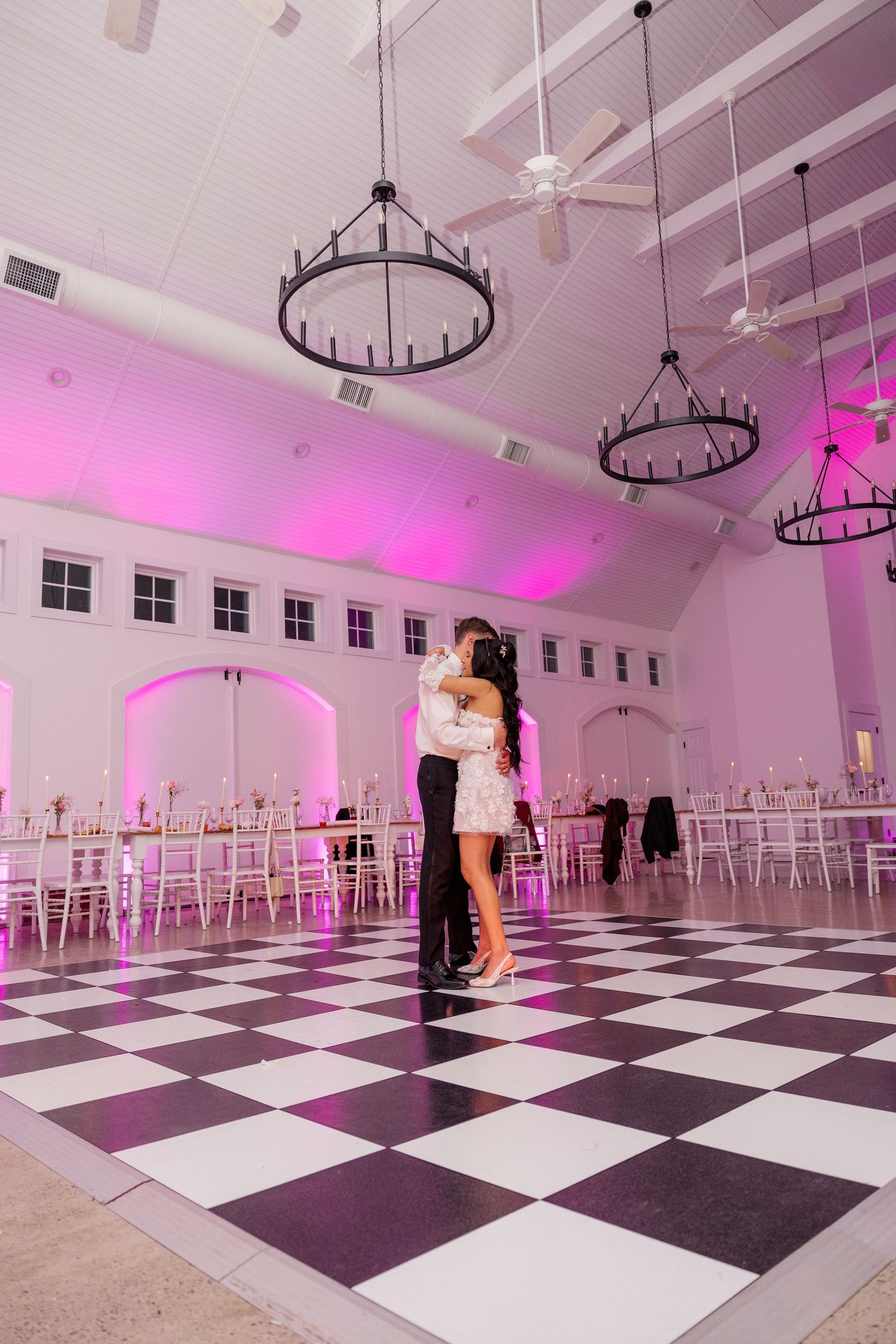 Couple sharing an intimate dance moment on a black and white checkered ballroom floor under chandeliers and pink lighting.