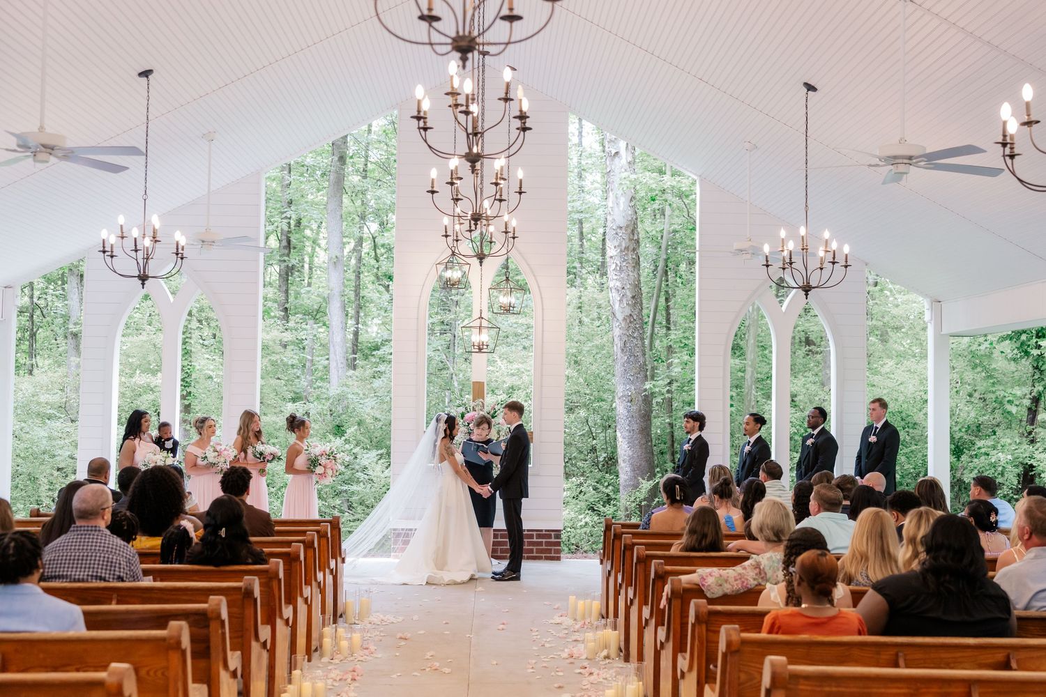A wedding ceremony in a bright chapel with tall windows and chandeliers surrounded by guests in pews.