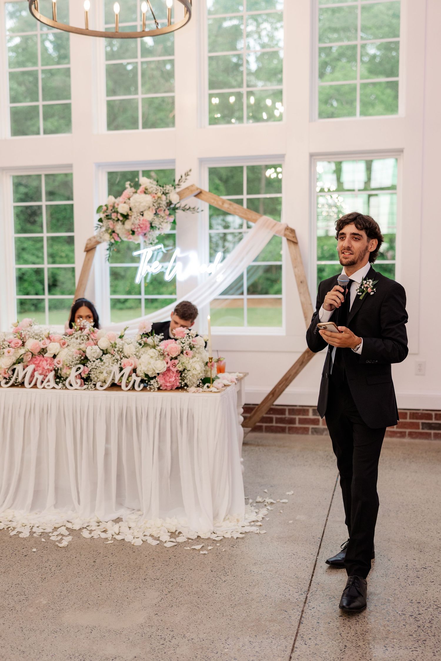 Wedding reception scene with speaker addressing guests near floral-decorated head table.