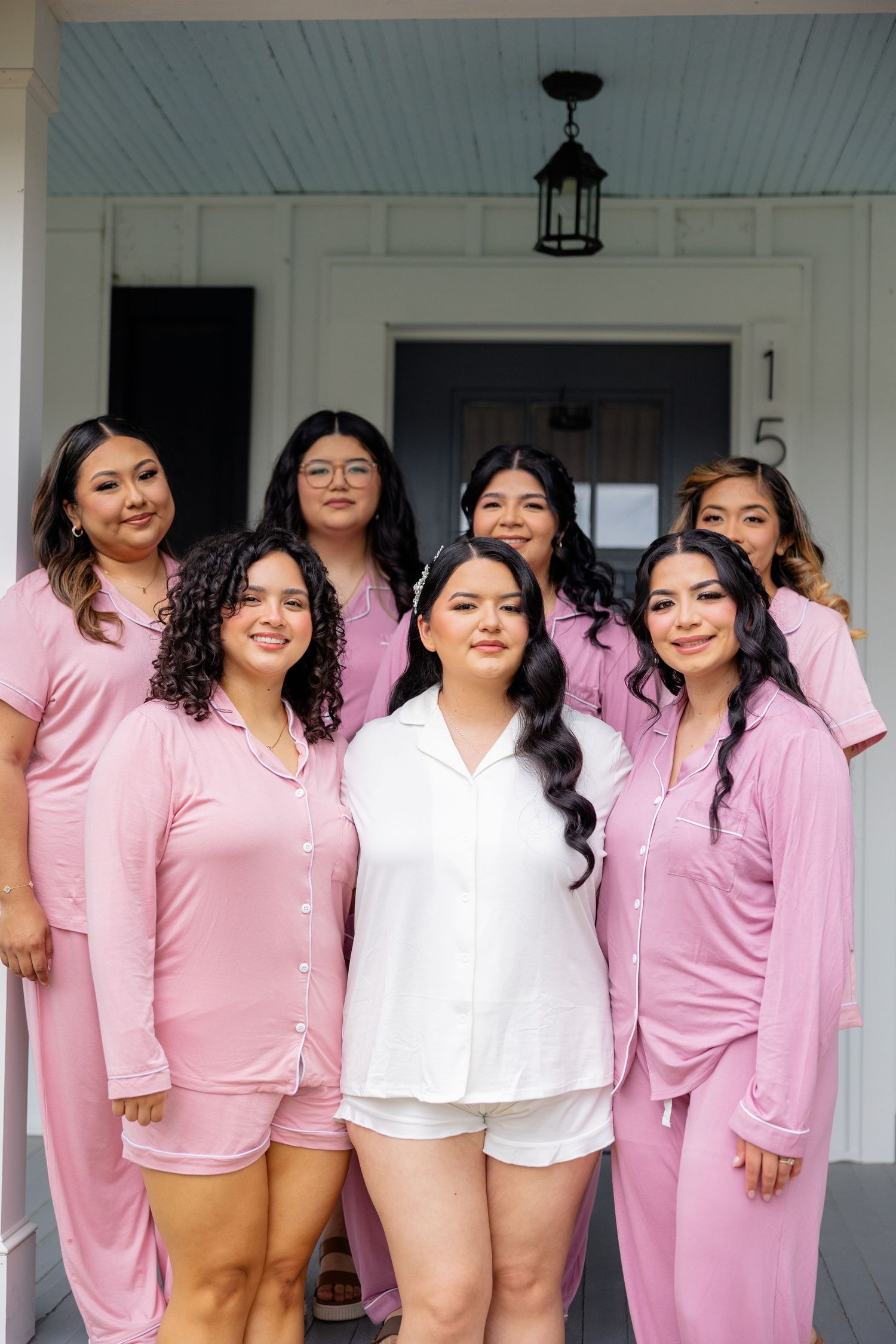 A group photo of bridesmaids wearing matching pink pajama sets posing together on a white porch.