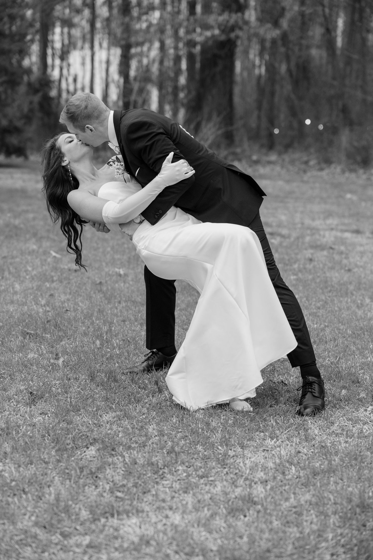 A romantic black and white photo of newlyweds sharing a dramatic dip pose outdoors in a wooded setting.
