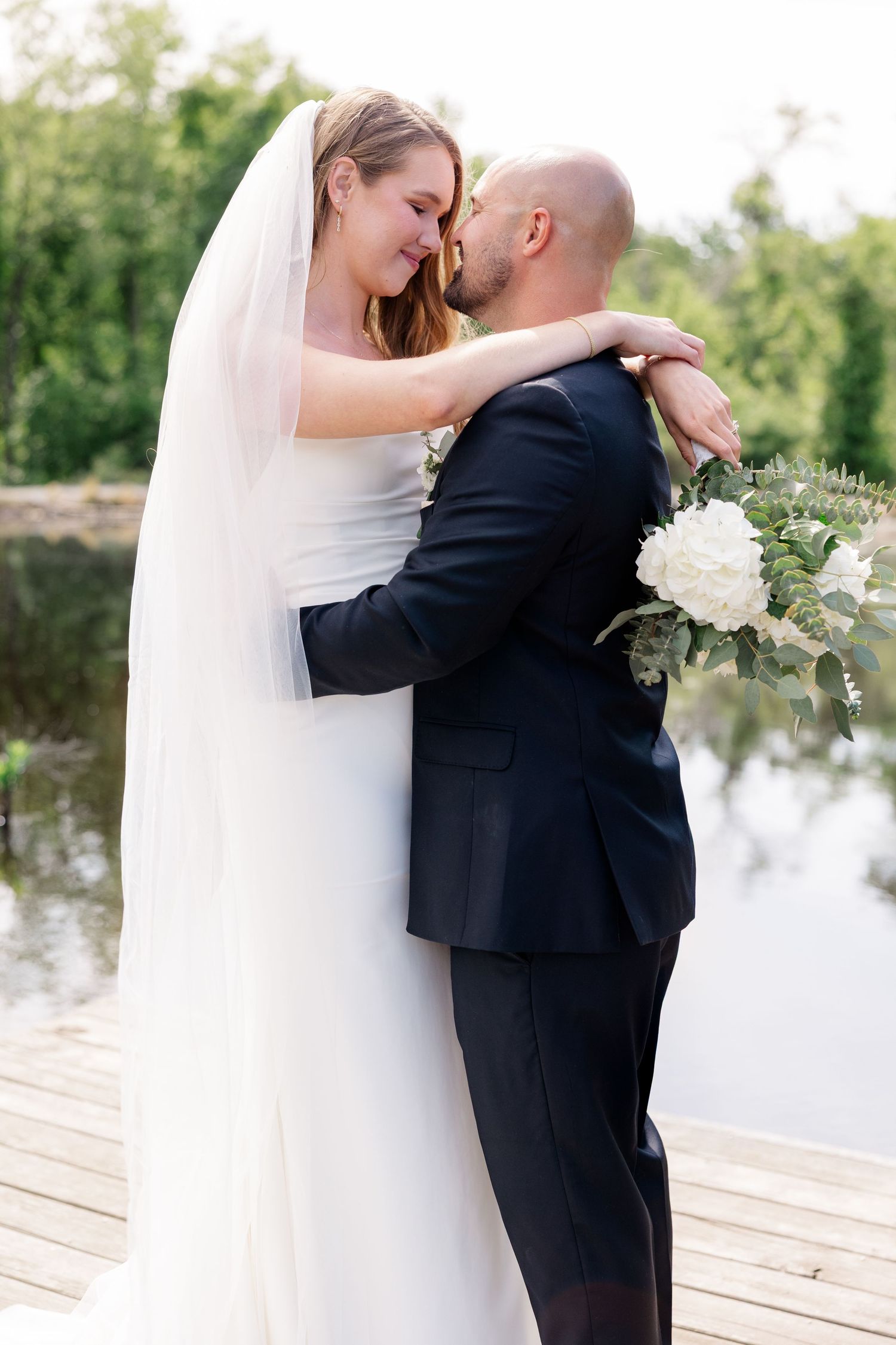 Romantic wedding couple embraces on a wooden dock overlooking a serene lake.