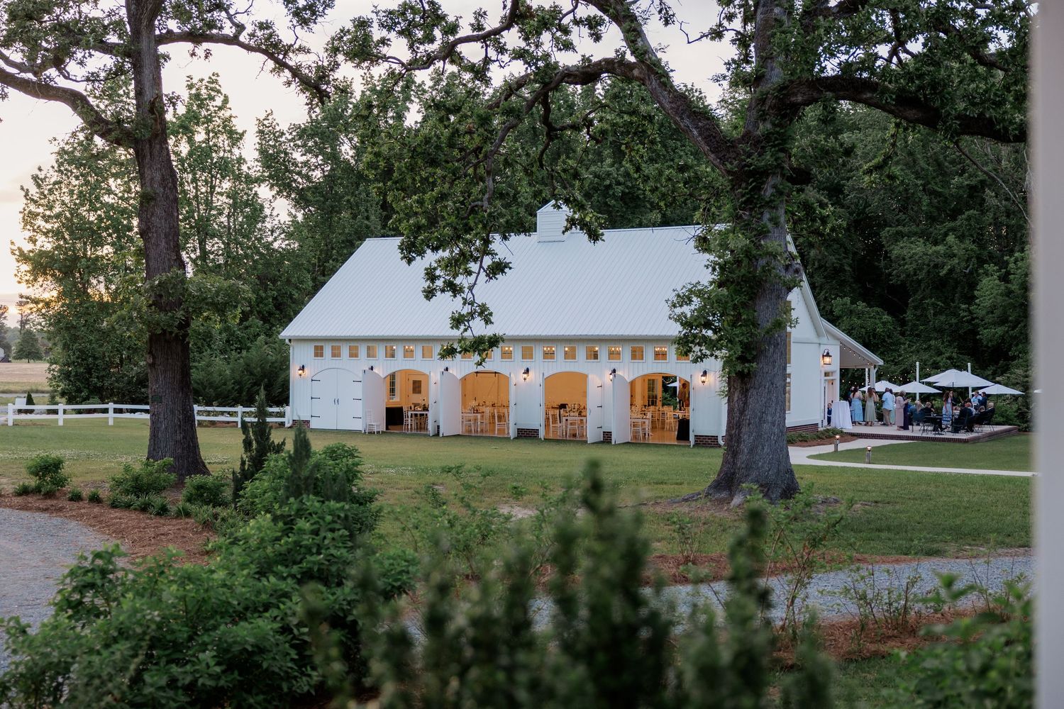 White farmhouse wedding venue surrounded by mature trees and green landscaping during evening golden hour.