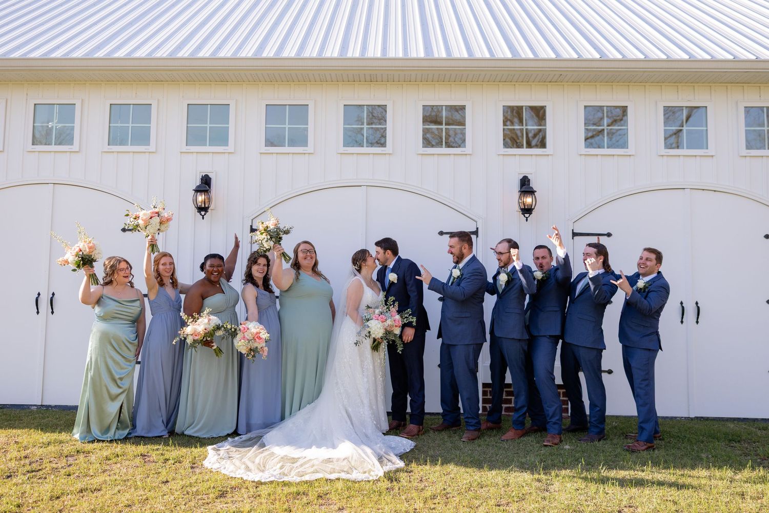 Wedding party in blue suits and sage bridesmaid dresses poses together in front of white barn doors.
