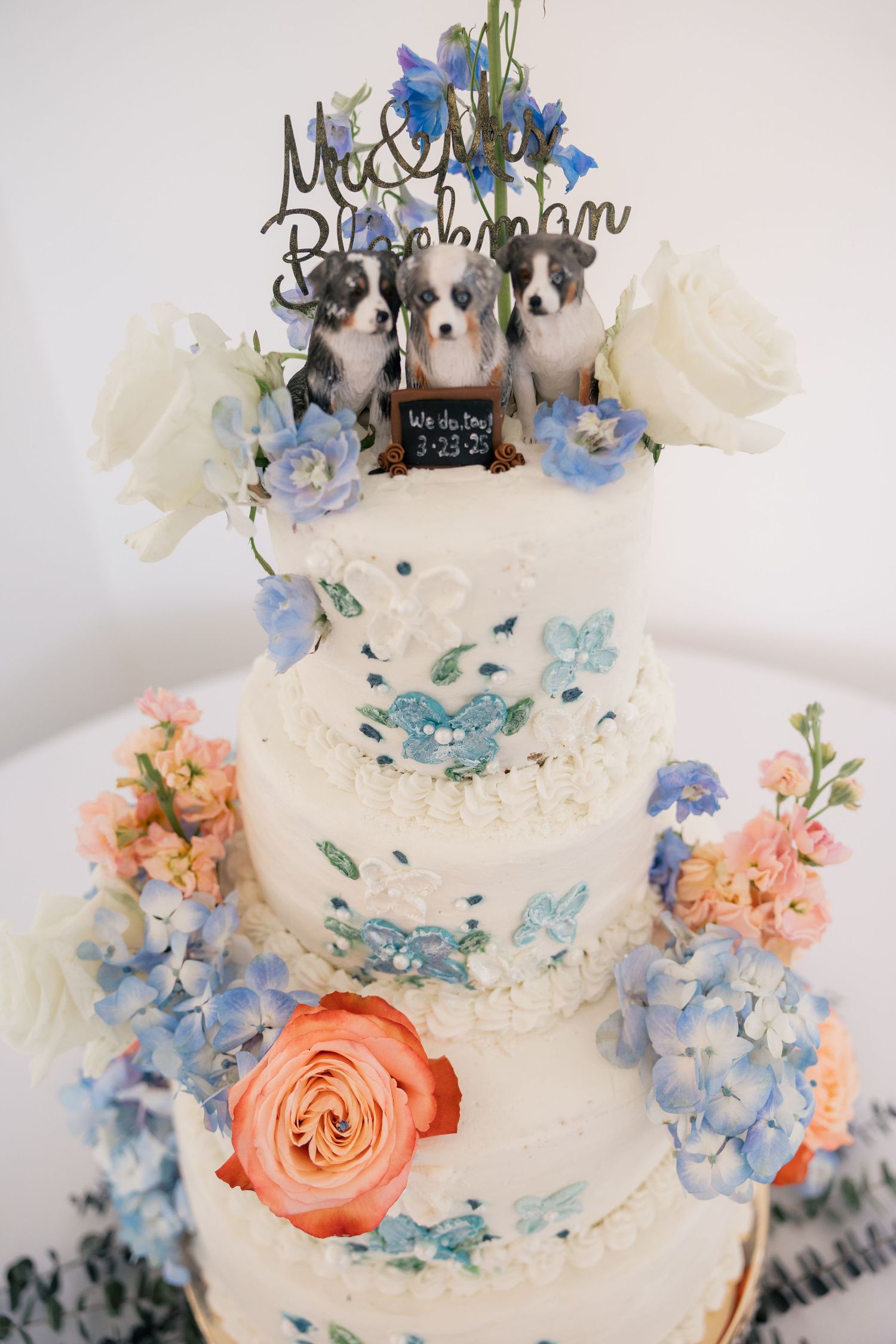 Three-tiered wedding cake decorated with blue and peach flowers and adorable dog figurines on top.