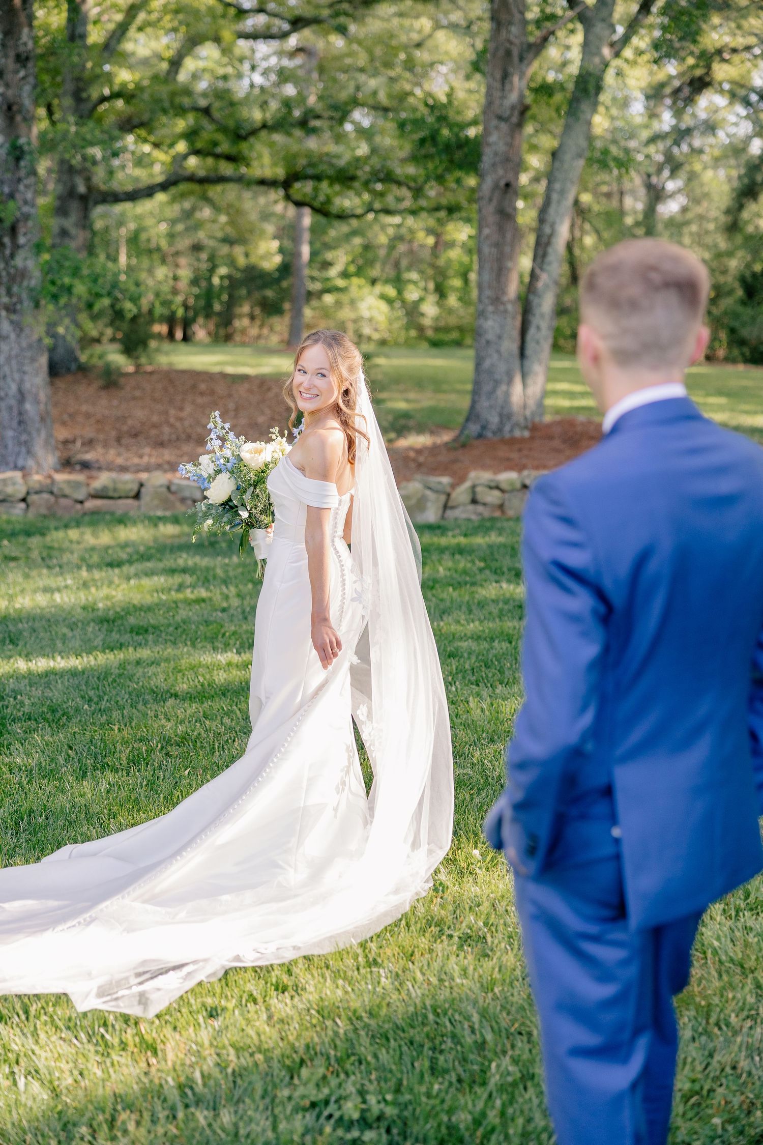 Bride in off-shoulder gown with long veil holds bouquet, smiling back at groom in blue suit on green lawn.