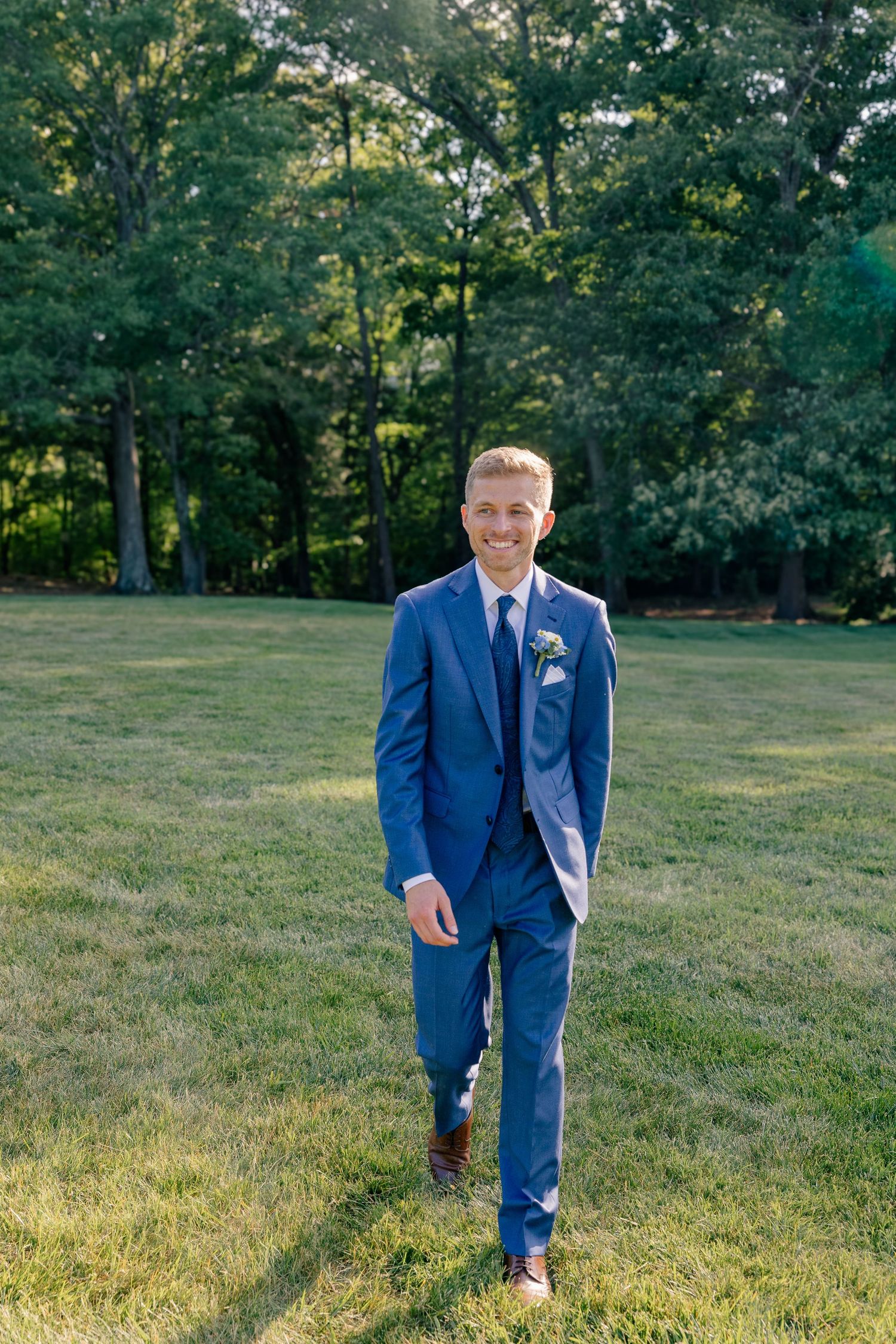 Groom in blue suit with boutonniere walks confidently across green lawn in golden sunlight.