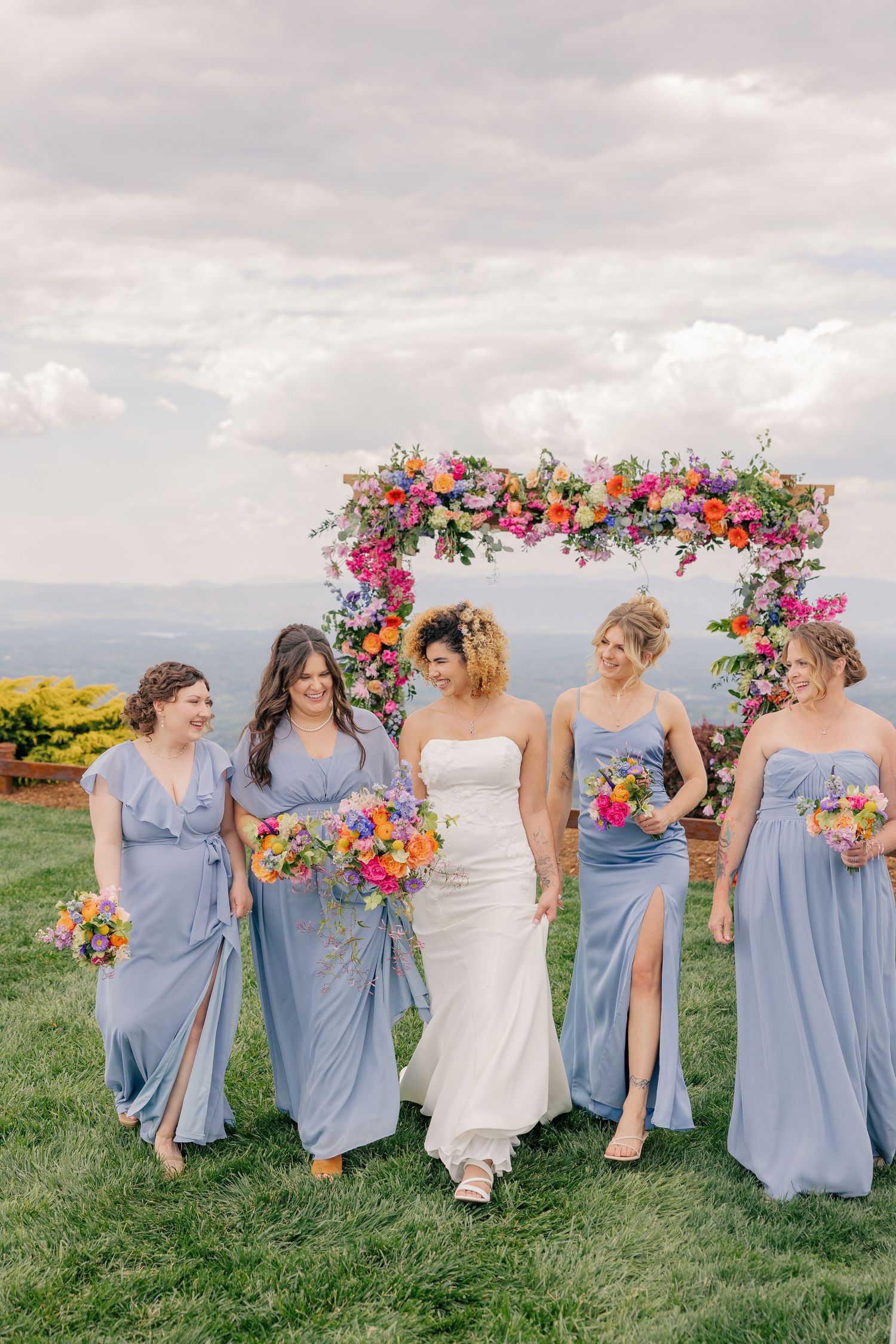 Bride in white gown walks with four bridesmaids in blue dresses before a colorful floral arch outdoors.