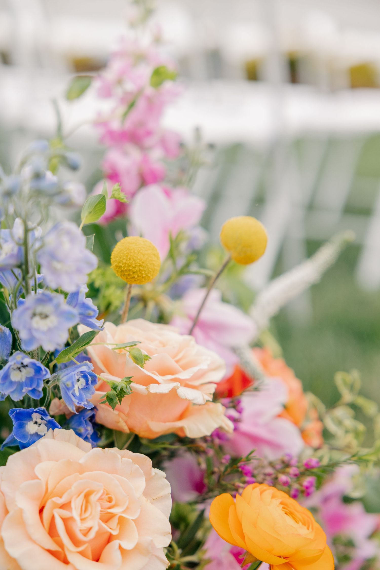 Colorful wedding floral arrangement featuring peach roses, blue delphiniums, yellow billy balls, and pink blooms outdoors.